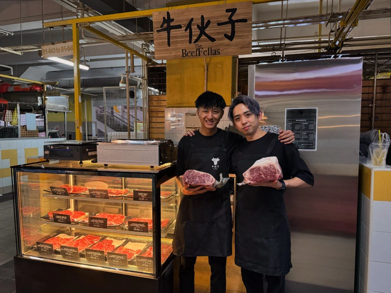 Two men standing in front of a beef display case, each holding a large cut of raw beef. They are inside a market or butcher shop, with a sign above that reads 'The Beef Fellas' in English and Chinese characters. Both are wearing black aprons.