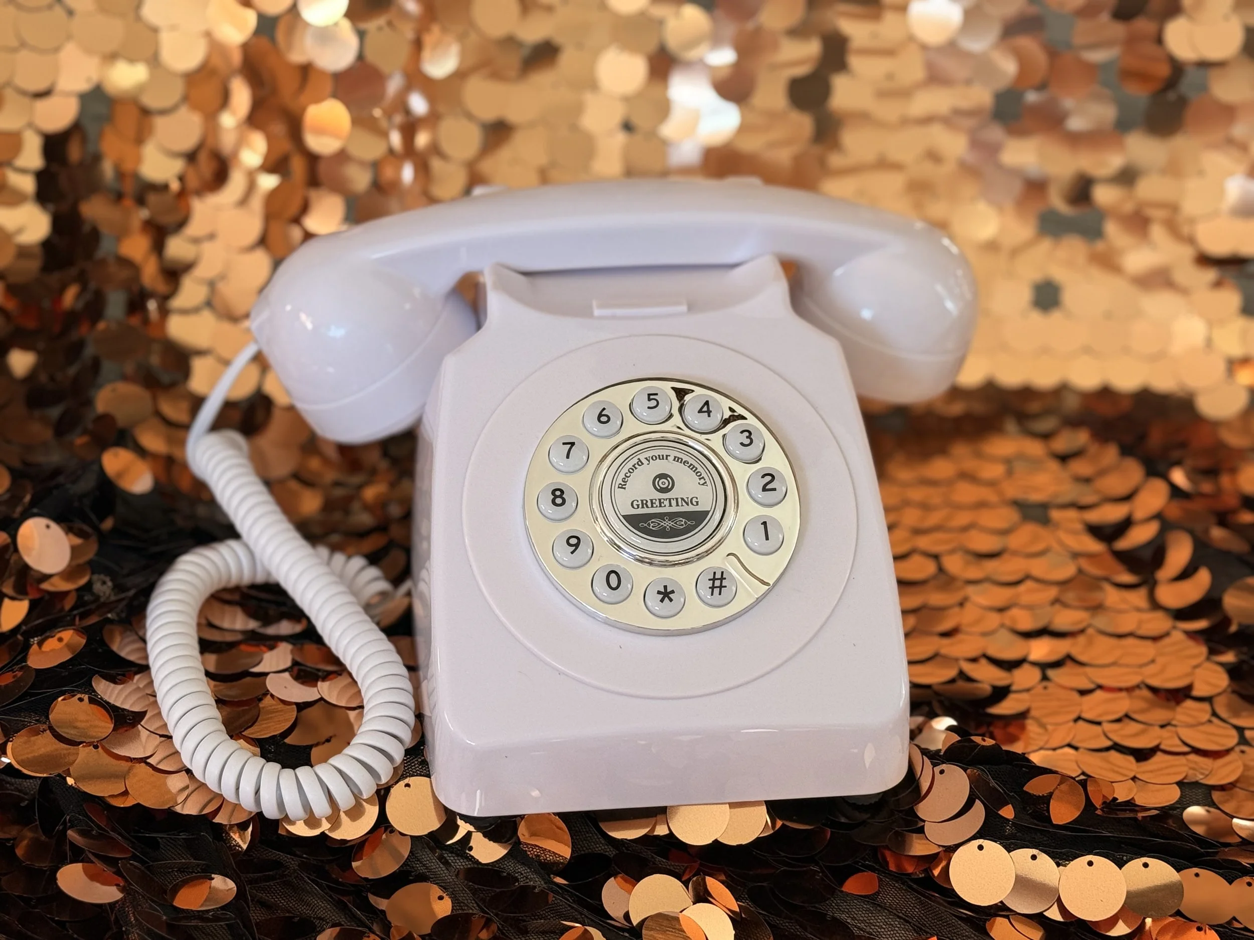 A white rotary telephone with a coiled cord, placed on a surface covered with orange and black sequined fabric.
