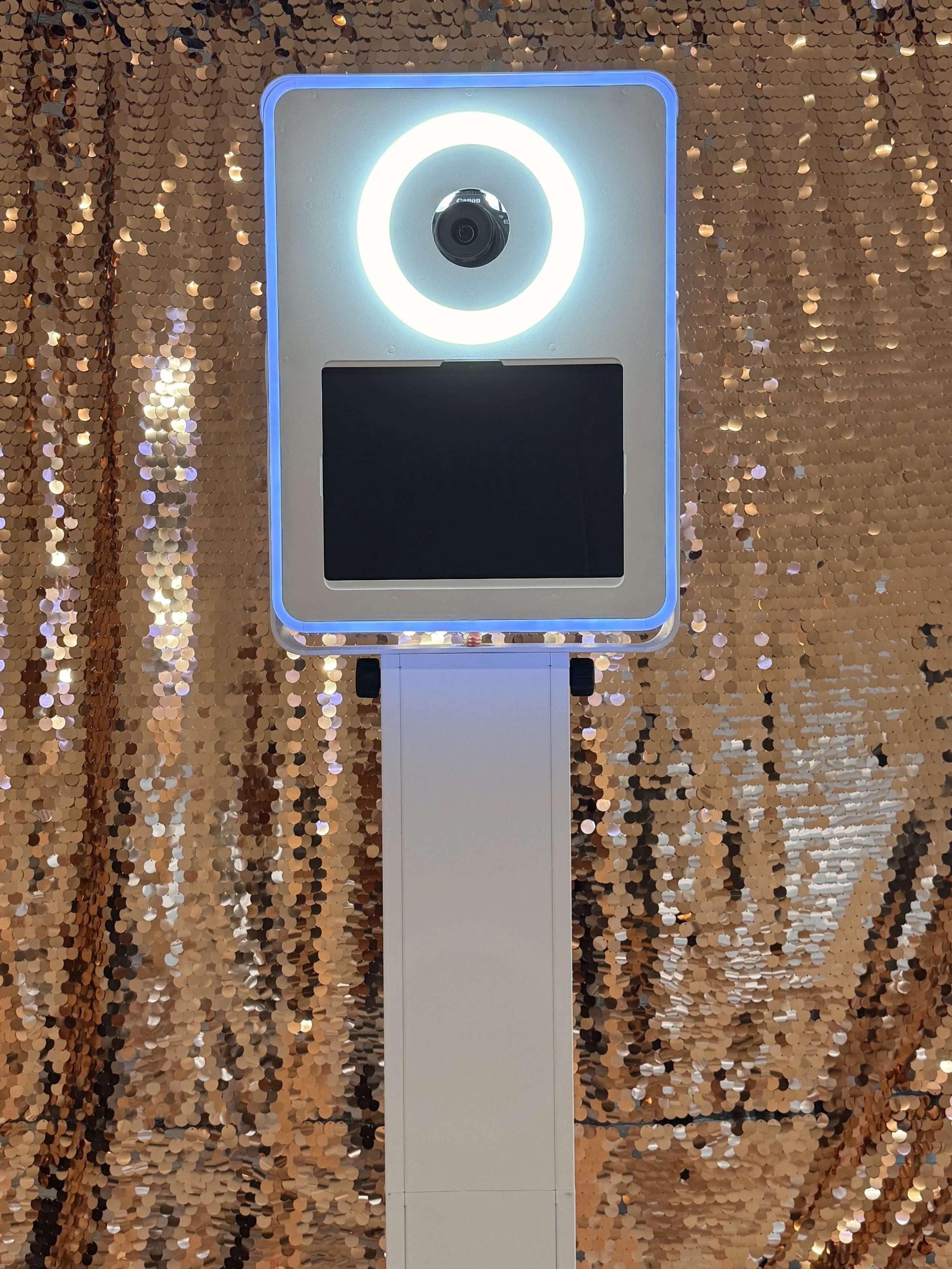 A photo booth with a circular ring light around the camera, positioned in front of a shiny, sequin-covered backdrop in gold and black colors.