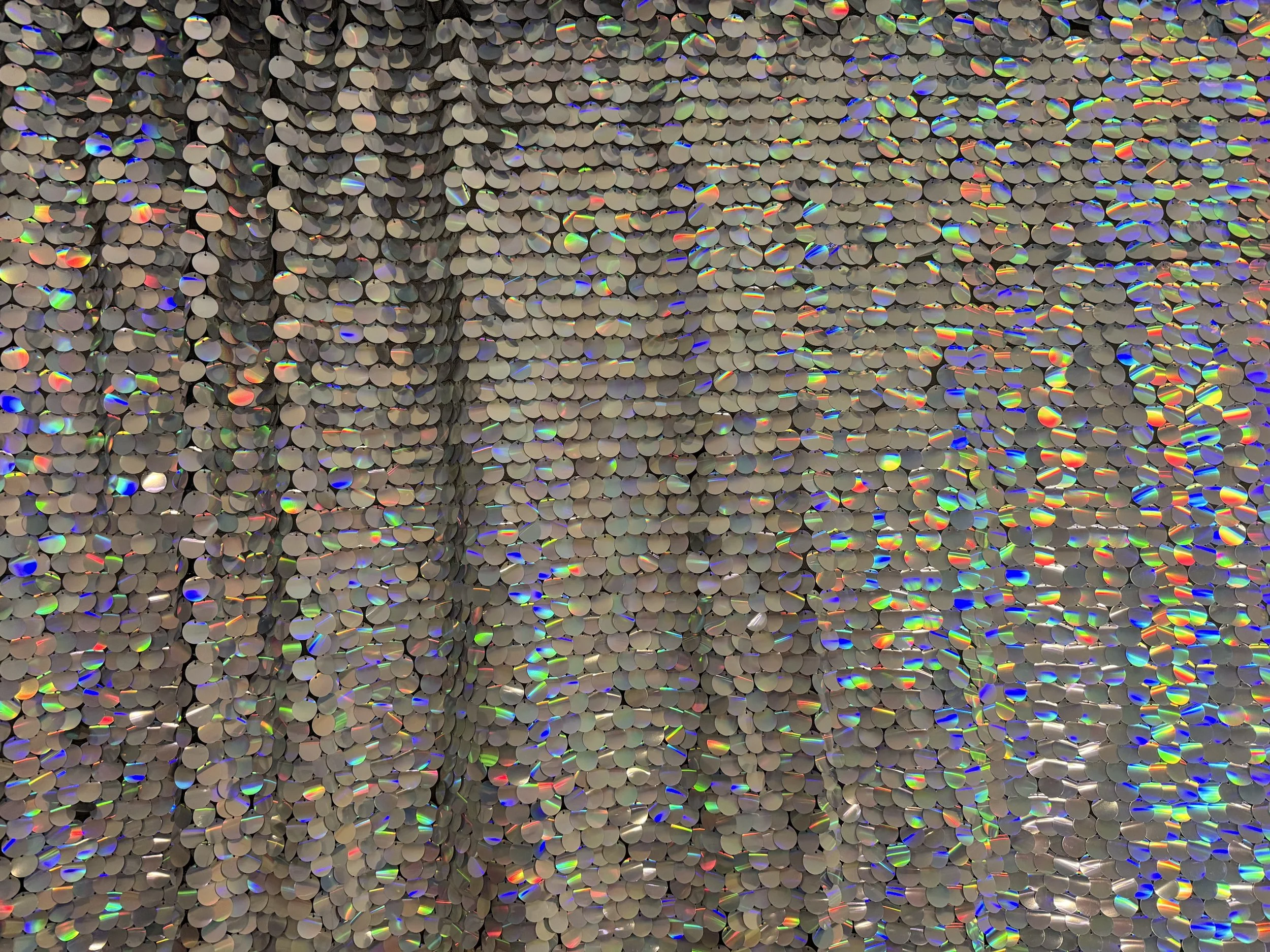 Close-up of a wall decorated with numerous small circular shiny discs that reflect light in rainbow colors. Unicorn silver iridescent sequin backdrop.
