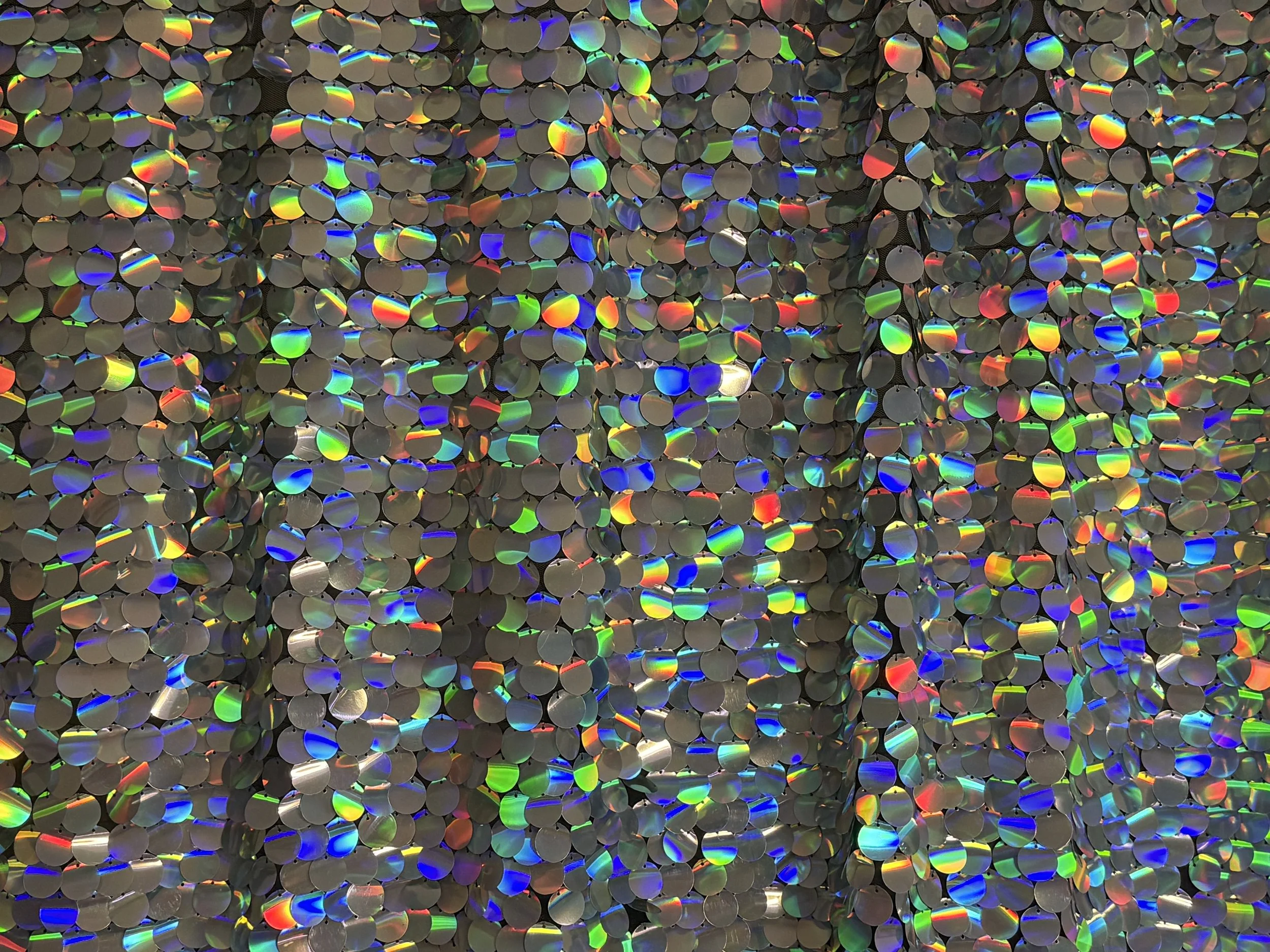Close-up of a decorative wall surface covered in small, circular, iridescent mirrors that reflect rainbow colors. Unicorn silver iridescent sequin backdrop.