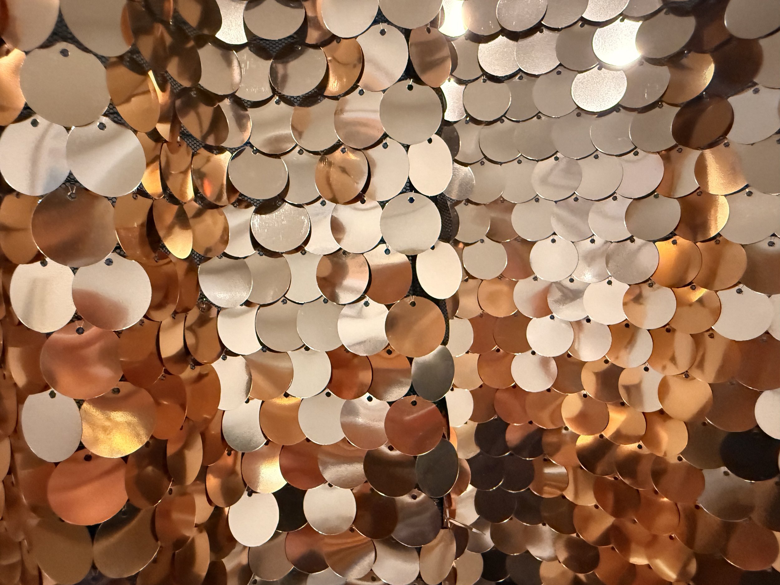 Champagne sequin backdrop. Close-up view of a decorative curtain made of numerous small, shiny, circular metallic discs in gold, copper, and silver colors, hanging closely together.