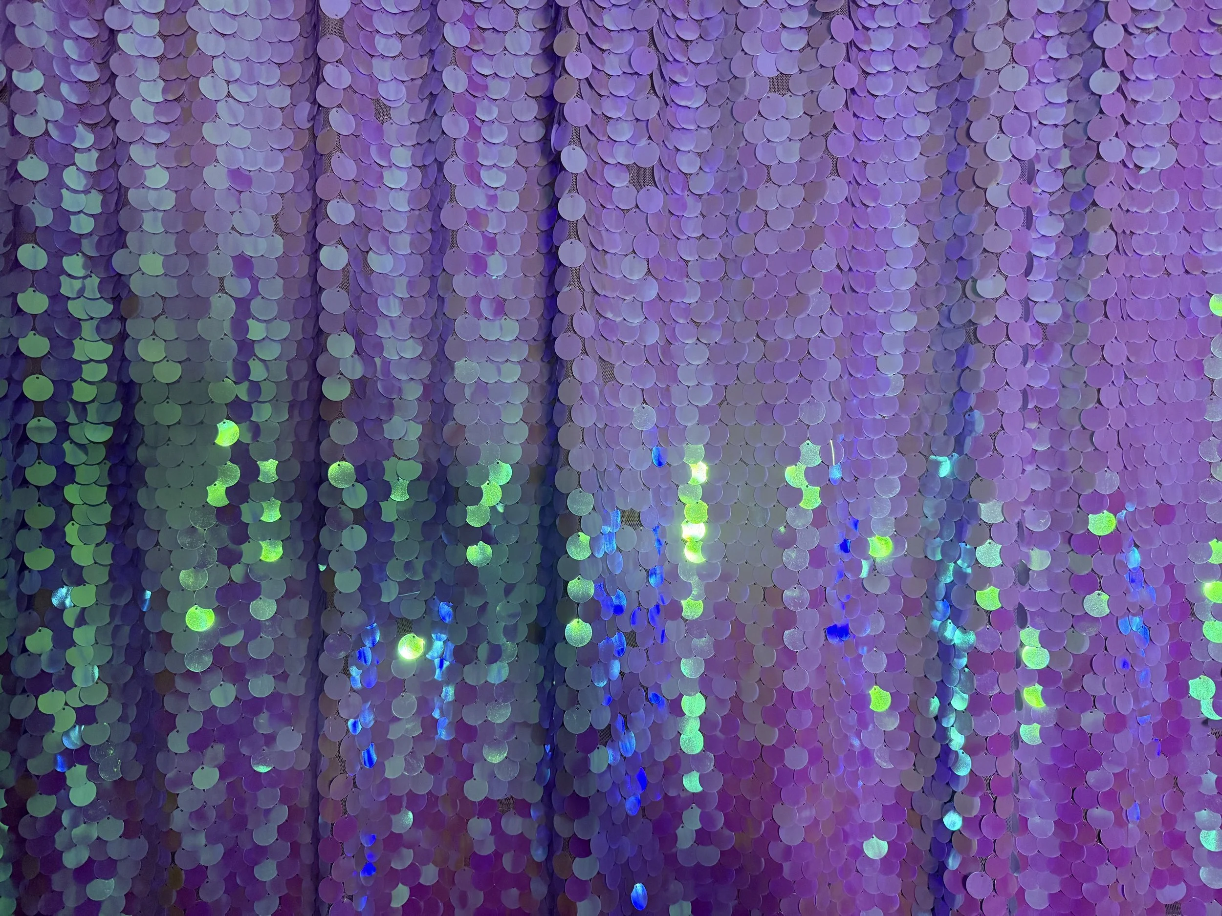 Close-up of a wall covered in purple, pink, and iridescent sequins or confetti, reflecting light in various colors.