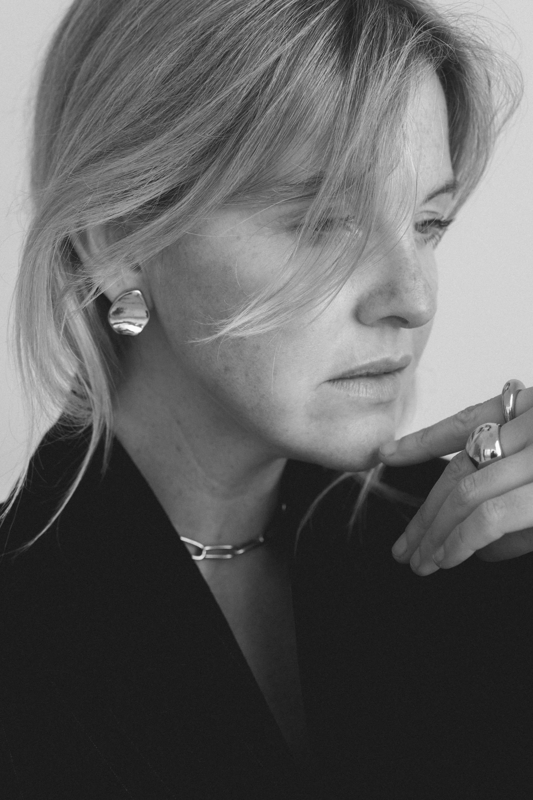 Schwarz-weiß-Fotografie einer nachdenklichen Frau mit kurzen, blonden Haaren, großen silbernen Ohrringen, einer Kette und mehreren Ringen, die ihre Hand an ihr Gesicht hält.