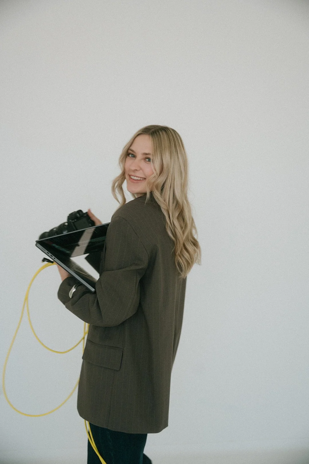 Eine junge Frau mit blonden, gewellten Haaren hält eine Kamera und ein Tablet in den Händen vor einem weißen Hintergrund. Sie trägt einen dunklen, langärmligen Blazer.