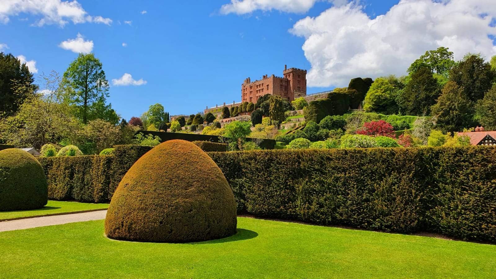 A scenic landscape with a historic castle on a hill, surrounded by lush trees and colorful bushes. There are manicured lawns and neatly trimmed hedges in the foreground.