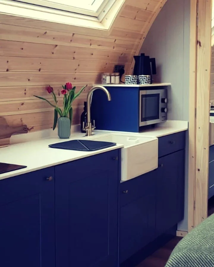 Small kitchen with blue cabinets, white countertop, a white farmhouse sink, a brass faucet, a vase with pink tulips, a microwave oven, a black coffee maker, and a skylight window in a wooden ceiling.