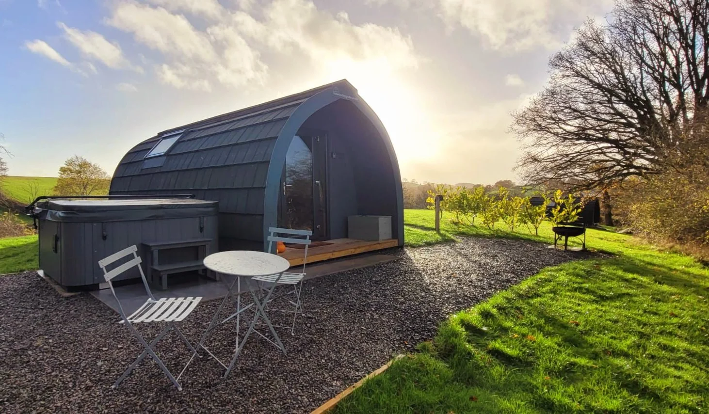 A Glamping pod in Wales set in a grassy field at sunset. There is a hot tub and a small outdoor table with two chairs on a gravel area, surrounded by trees and open landscape.