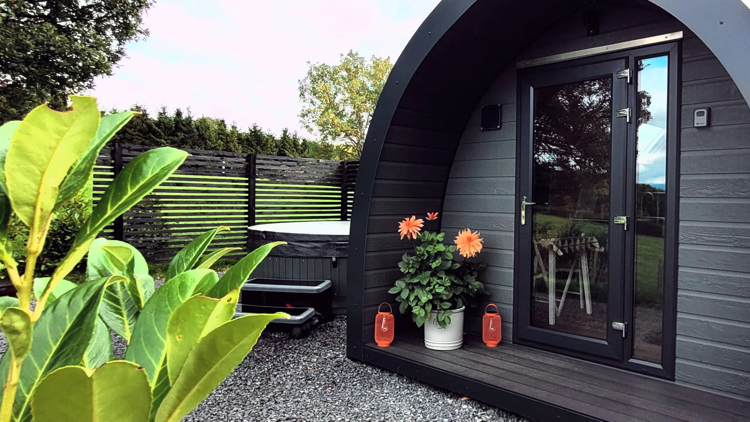 An Glamping pod in the Welsh countryside with potted flowers and lanterns. There is a hot tub and a wooden privacy fence in the background, surrounded by greenery and trees.