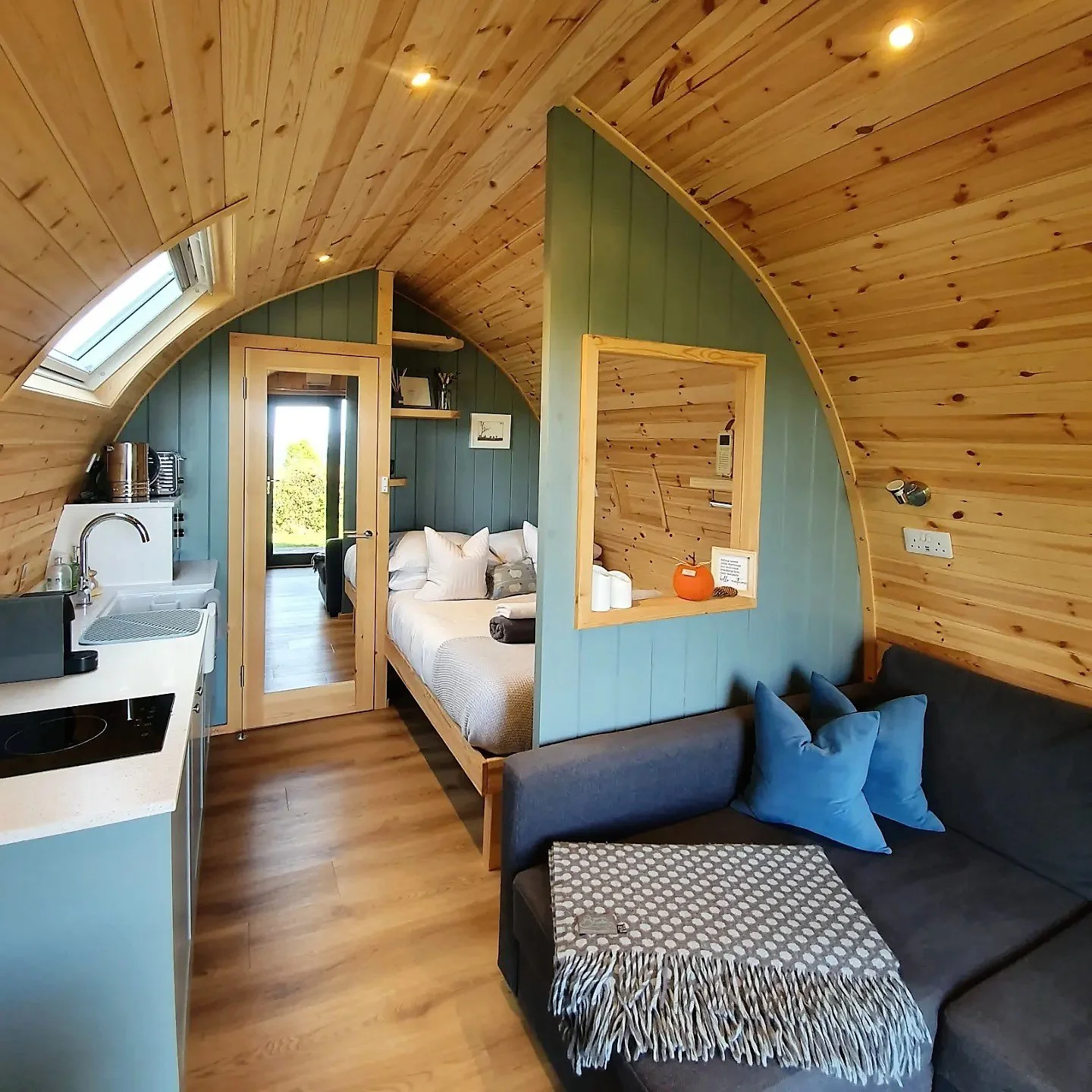 A cozy tiny home interior with wooden curved ceiling, a small kitchen area with a sink and stovetop, a hallway leading to a bedroom with a bed and pillows, a window with a view, and a sitting area with a dark sofa, blue cushions, and a patterned blanket.