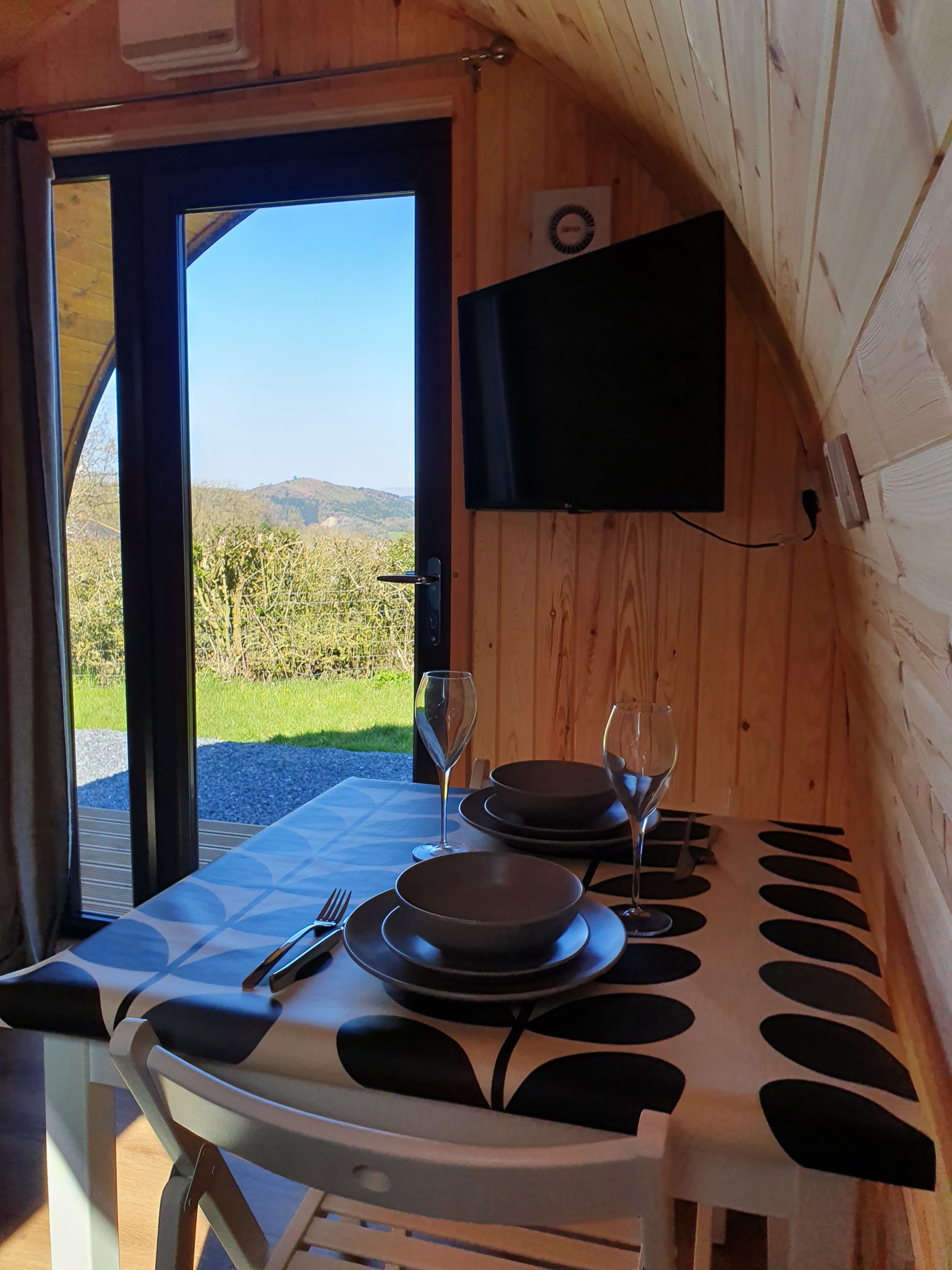 Indoor dining area with a table set for two, featuring Orla Kierly tablecloth, bowls, plates, utensils, and glasses, near glass door leading to a green outdoor landscape with hills and trees.