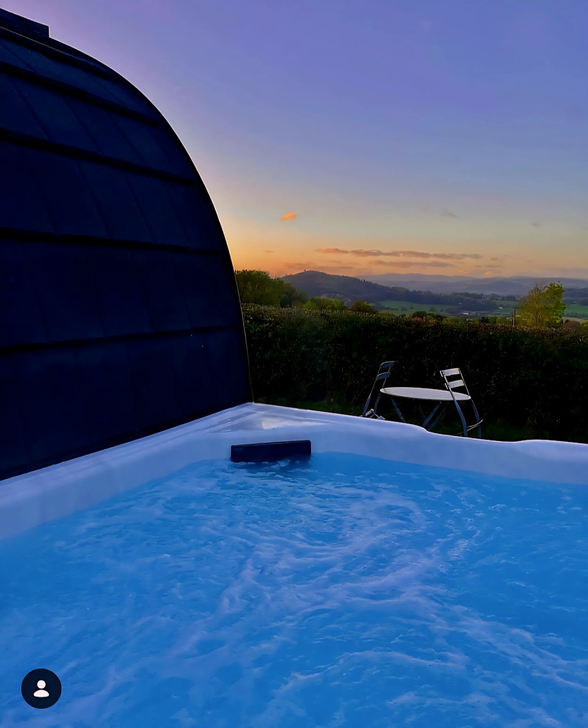 Hot tub with bubbling water overlooking a scenic landscape at sunset, with green hills and a clear sky.