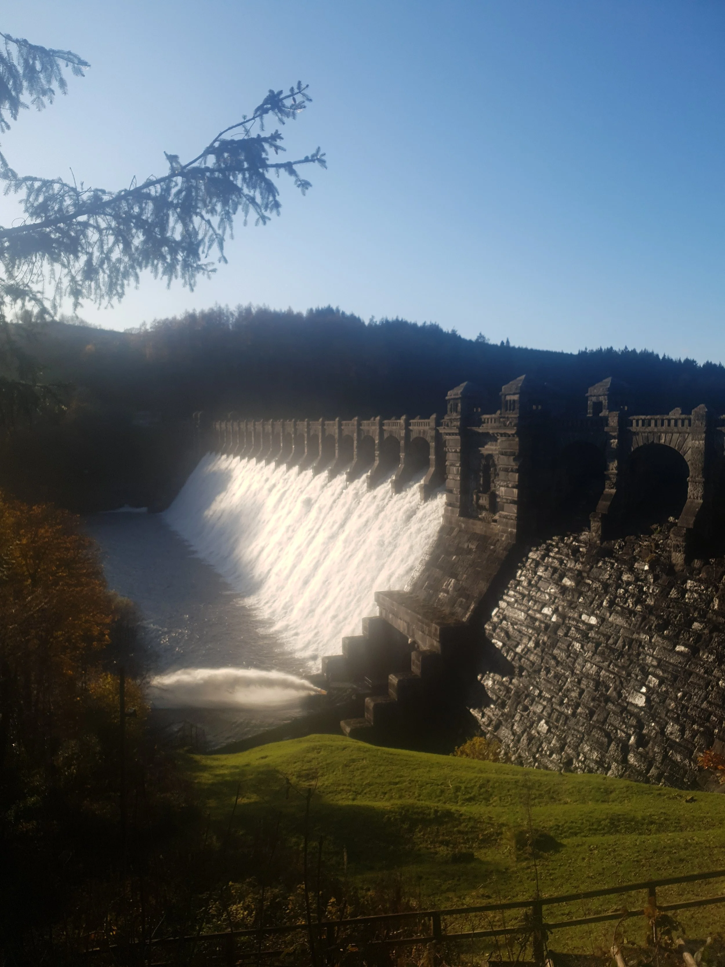 A dam with water spilling over the top, surrounded by trees and hills under a clear blue sky.