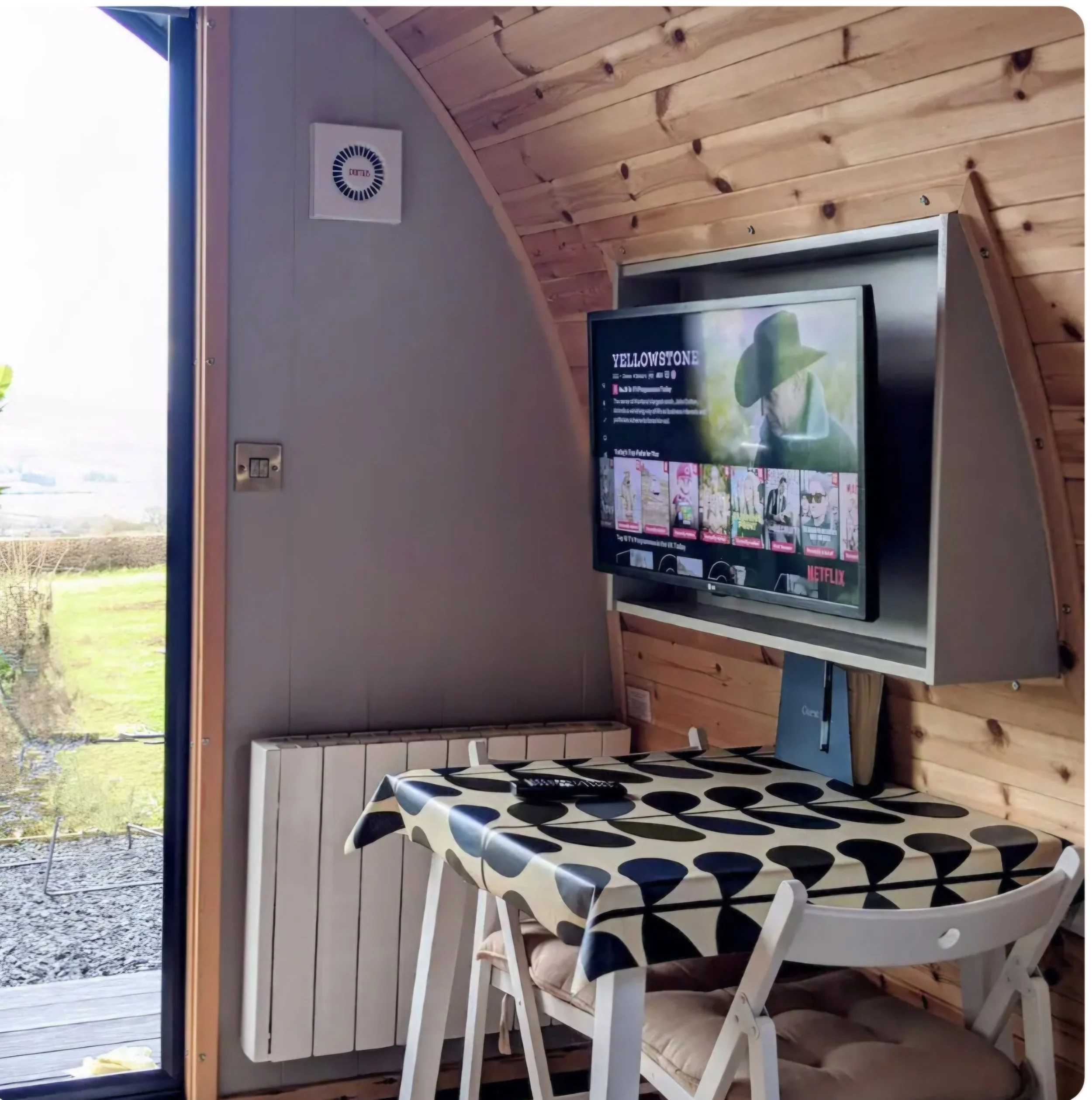 Interior of a small wooden cabin with a table covered with a Orla Kierly polka dot tablecloth, a Smart TV mounted on the wall displaying Netflix, a remote control on the table, and a window showing the rolling fields and countryside view. 