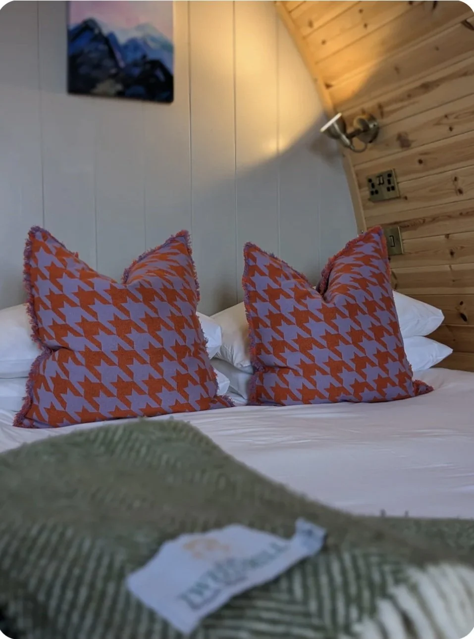 A cozy bedroom with a bed featuring Tbco patterned pillows, a green British wool blanket, white sheets, a wooden wall with a sconce light, a USB power outlet, and a landscape painting on the wall.