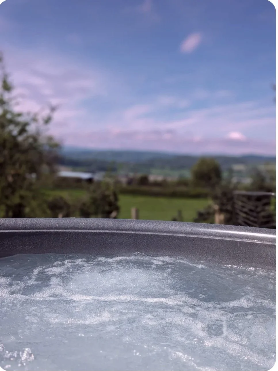 A hot tub outdoors with a view of green fields, trees, and a blue sky with clouds in the background.