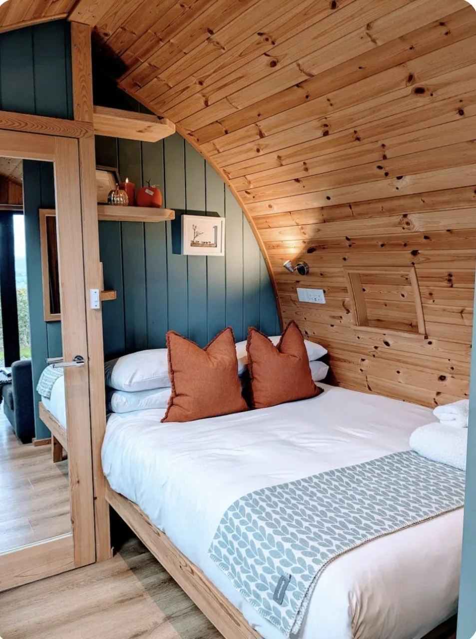 A cozy bedroom with a wooden sloped ceiling and a bed with white linens, two orange pillows, and a decorative gray and white blanket.