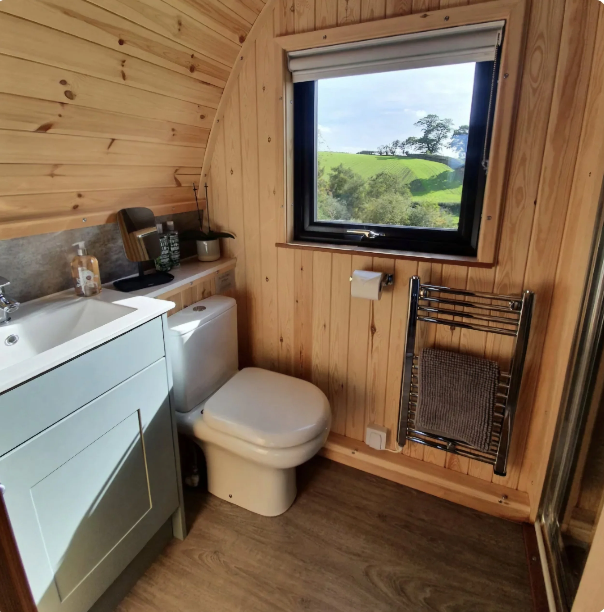 Bathroom with wooden walls, a window showing a scenic green landscape, a white toilet, a sink with a soap dispenser, a small mirror, a towel rack with a towel rail & shower. 