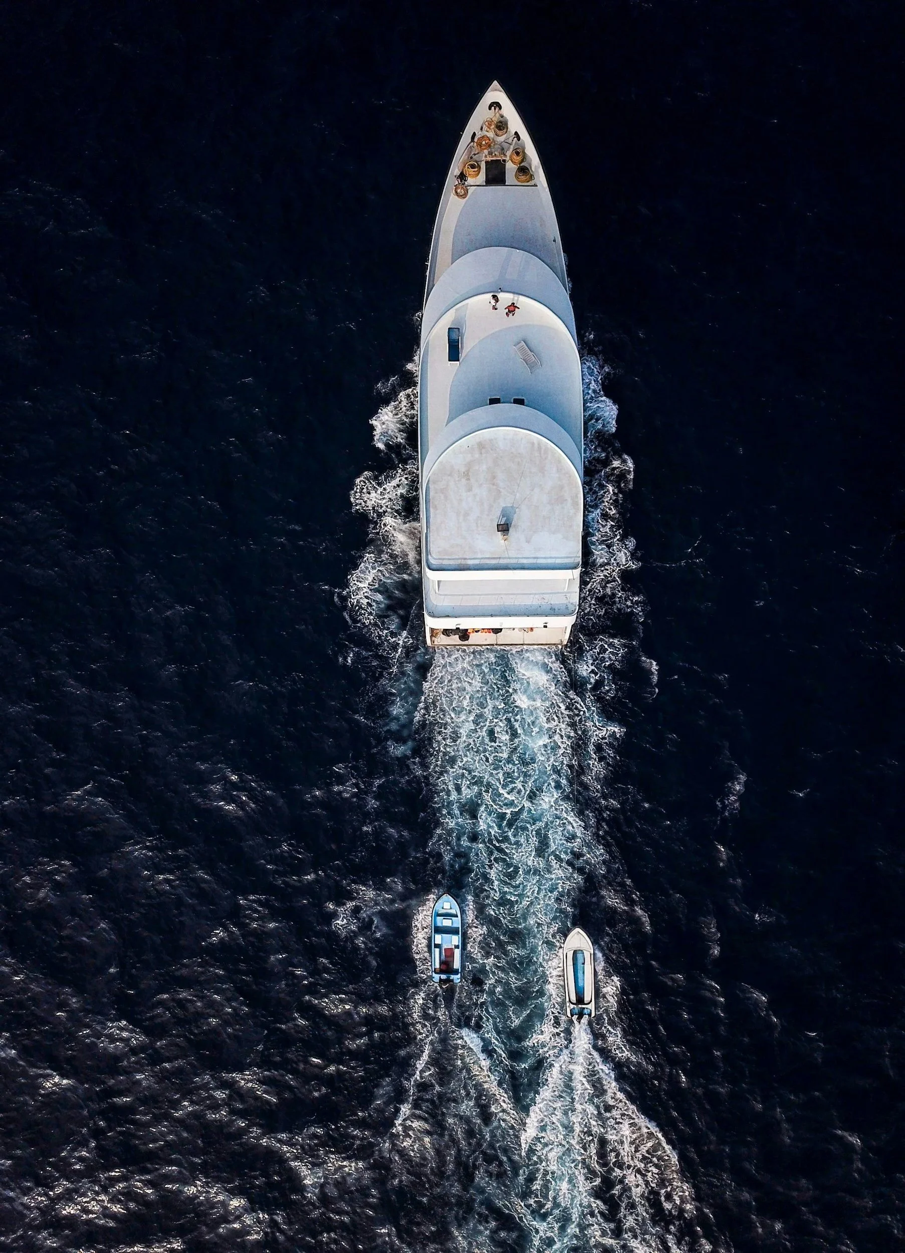 Eine große weiße Yacht fährt auf dem Meer, begleitet von zwei kleineren Booten. Auf der Yacht sind mehrere Personen sichtbar, die auf dem Deck stehen oder sitzen.