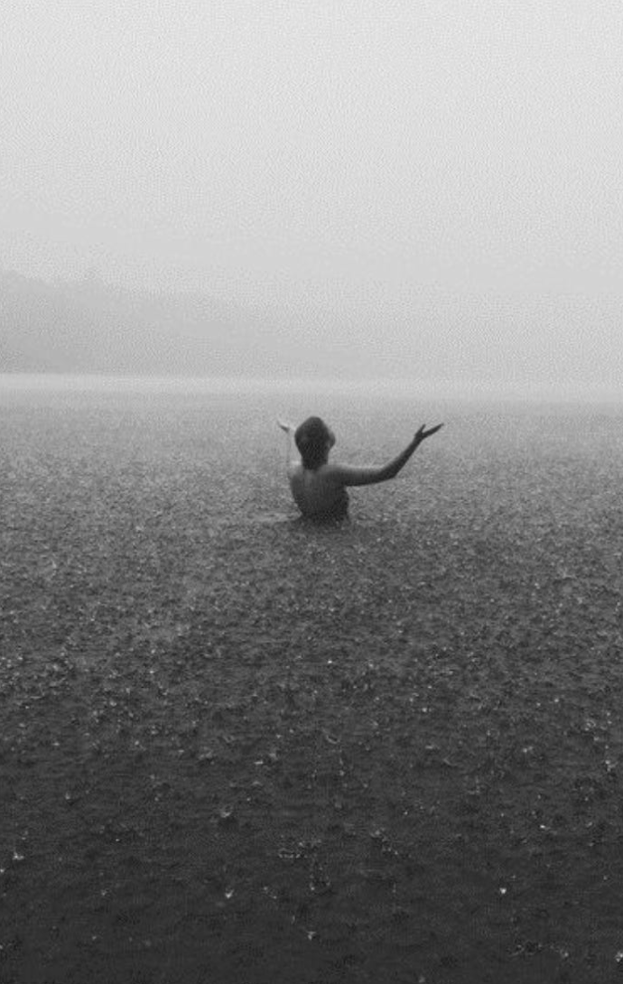 Une personne, vue de dos, émergent de la mer, avec les bras levés, dans un paysage en noir et blanc.