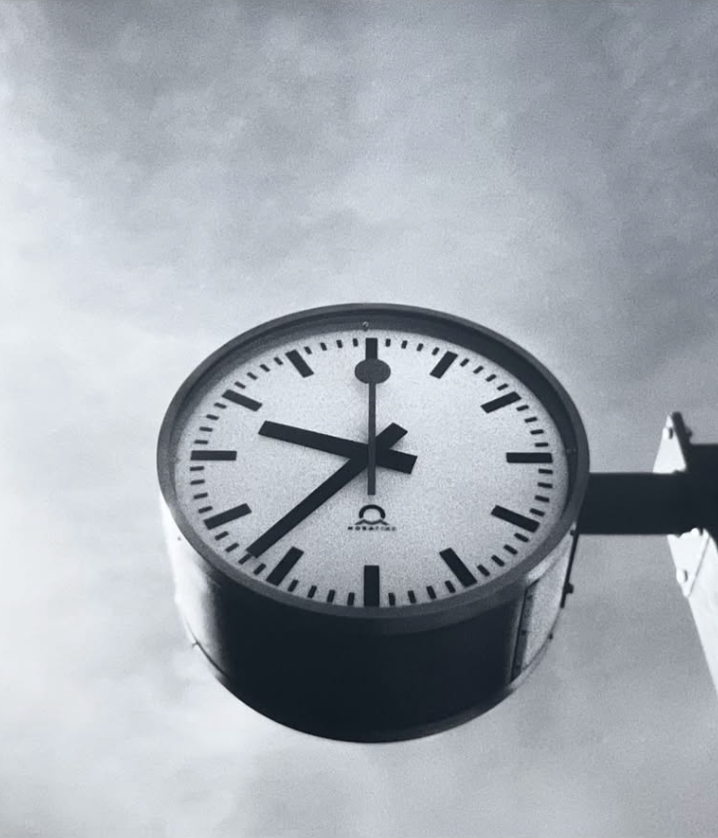 Horloge d'arrêt avec un cadran blanc, chiffres noirs, et aiguilles indiquant environ 7h41, fixée à un support contre un ciel nuageux.