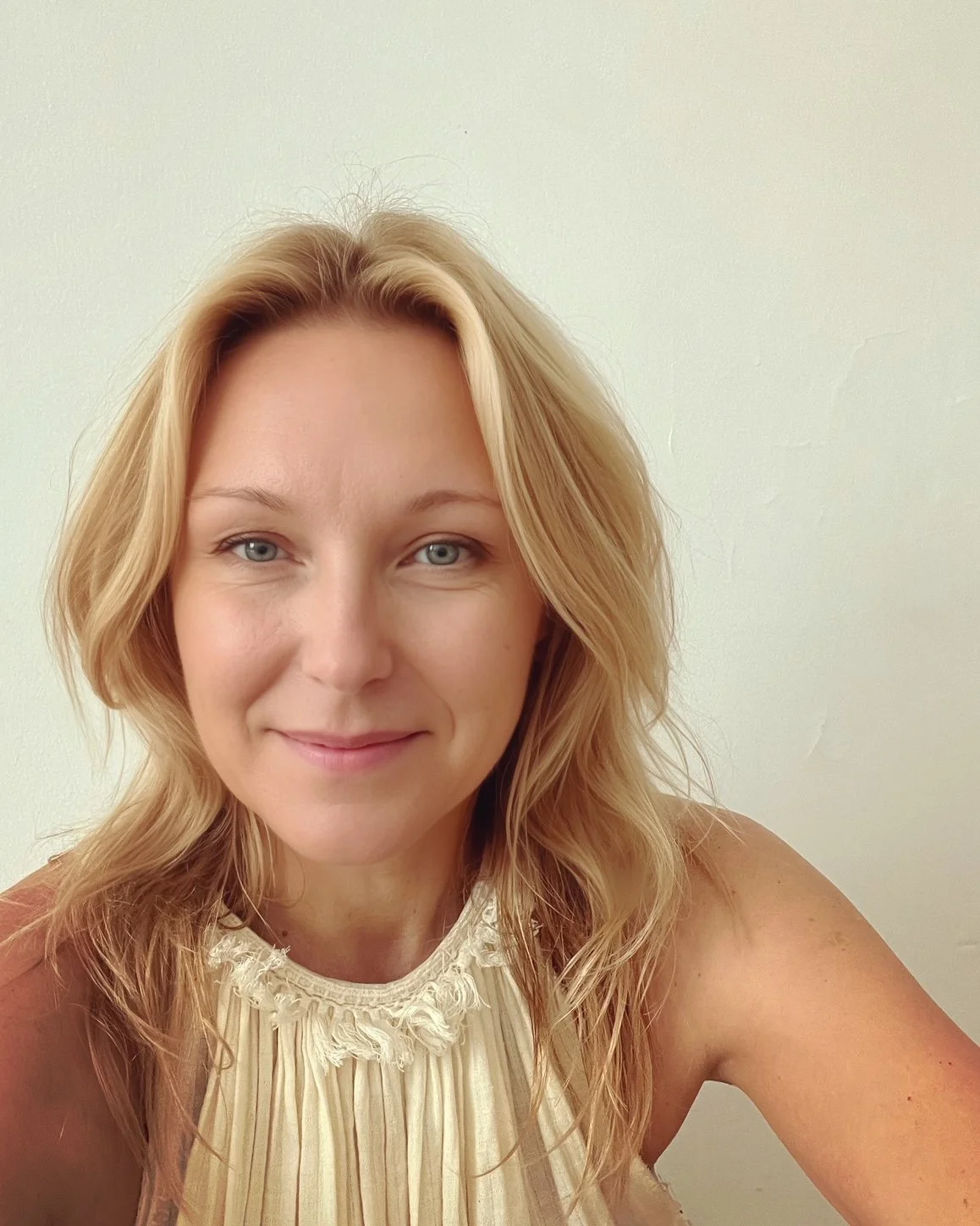 Un portrait d'une femme aux cheveux blonds, souriante, portant une robe crème avec des détails en tulle, devant un mur blanc.