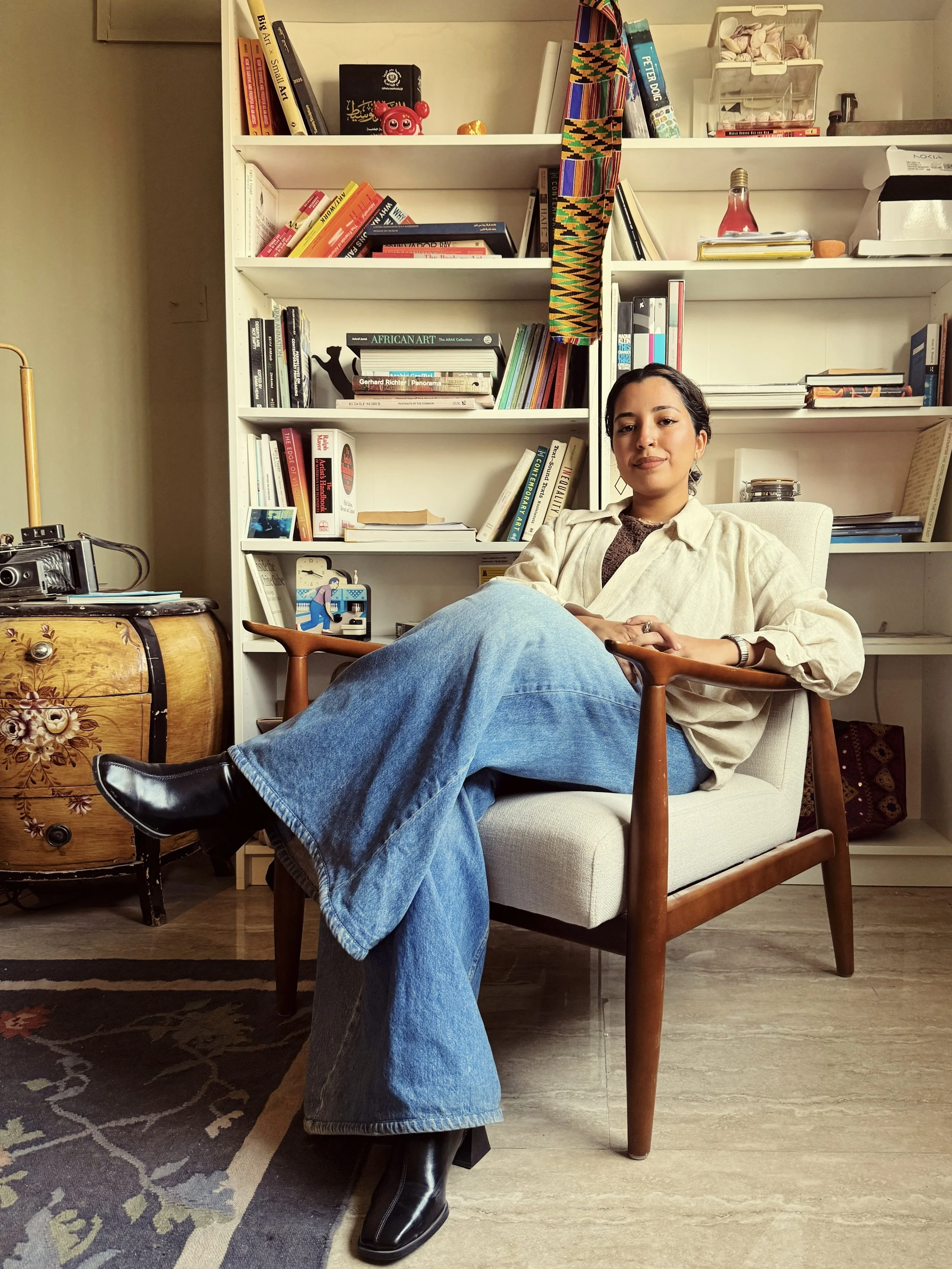 A woman sitting comfortably in a modern armchair with crossed legs, wearing a beige shirt, blue jeans, and black boots, in a room with a white bookshelf filled with books and decorative items behind her, and a vintage wooden chest beside her.