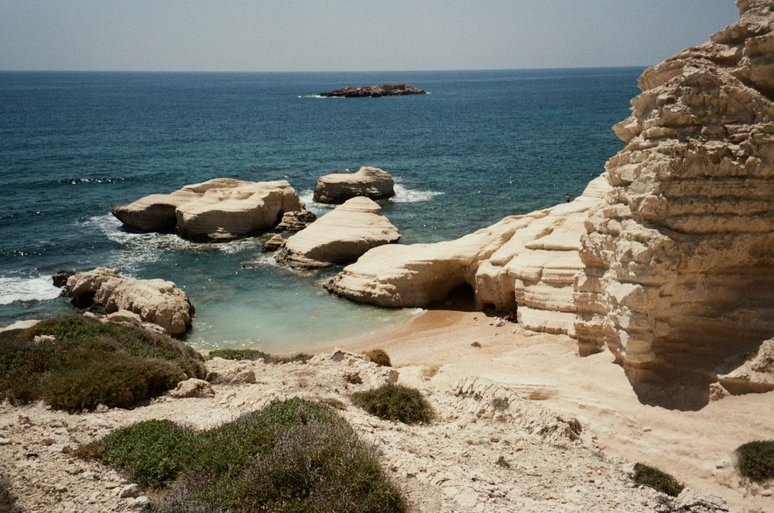 Sunny beach with large white rocks, sandy shore, and desert vegetation in the foreground, ocean with small island in the background.