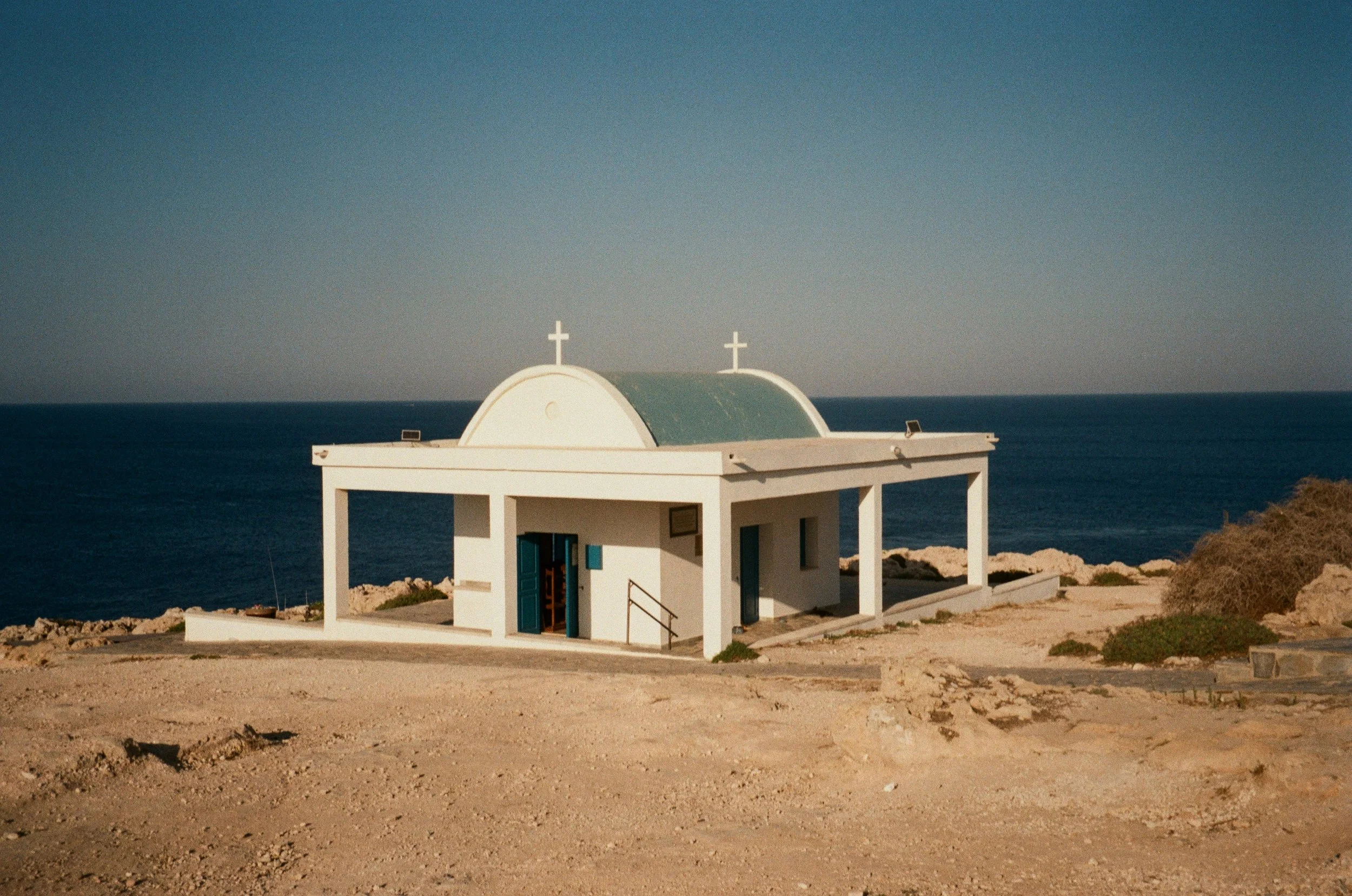 Small white chapel with blue doors and crosses on top, situated on a rocky beach overlooking the ocean under a clear sky.
