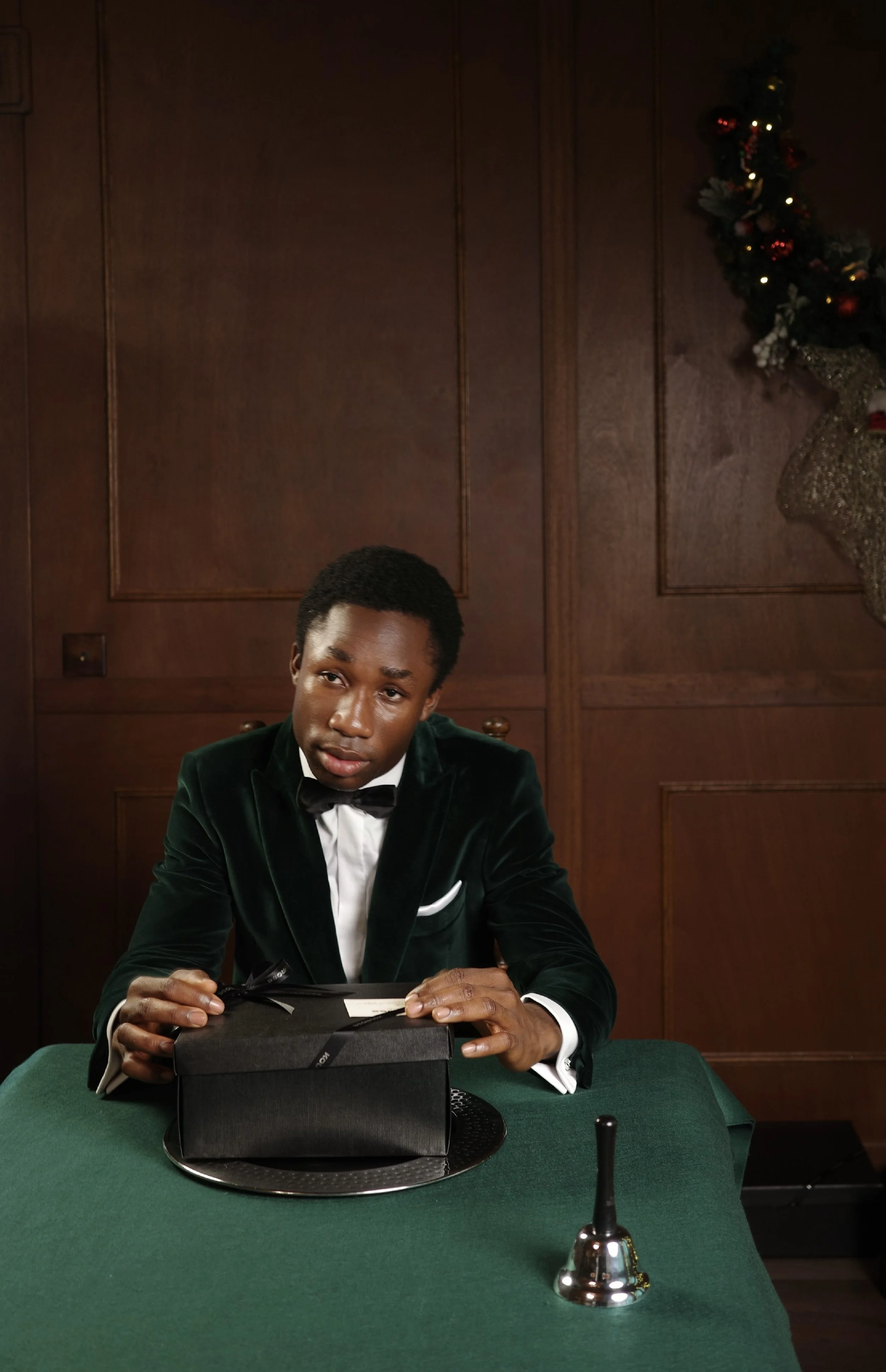 A young man in a tuxedo with a velvet blazer, sitting at a green table with a black gift box, in a wood-paneled room decorated for Christmas.
