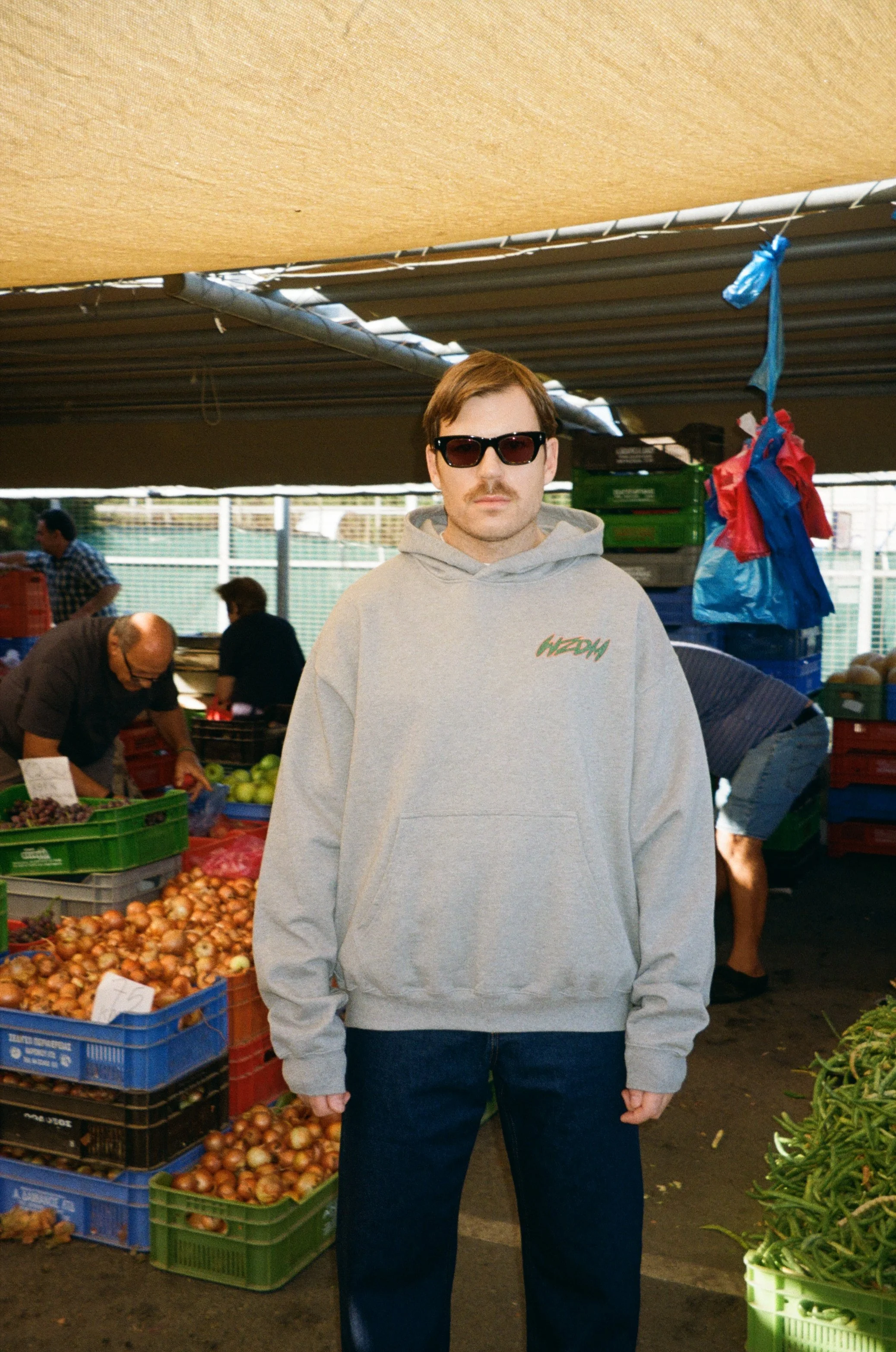 A man wearing sunglasses and a gray hoodie with green text stands at a produce stand at an outdoor market.