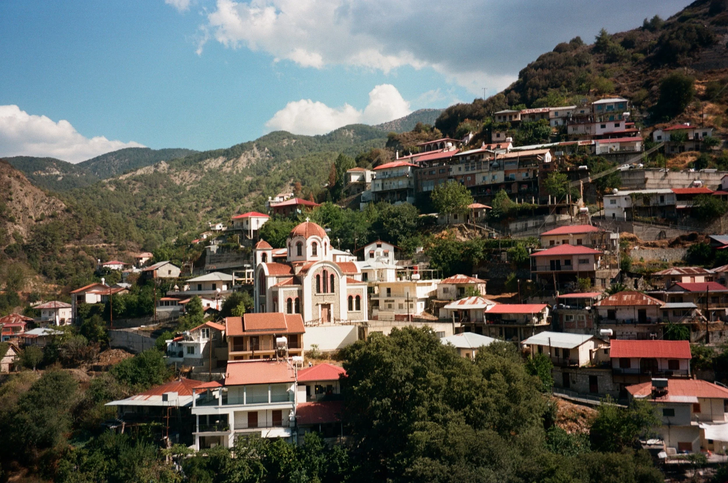 Hillside town with numerous white and beige houses, many with red roofs, surrounding a central church with a dome, under a partly cloudy sky.