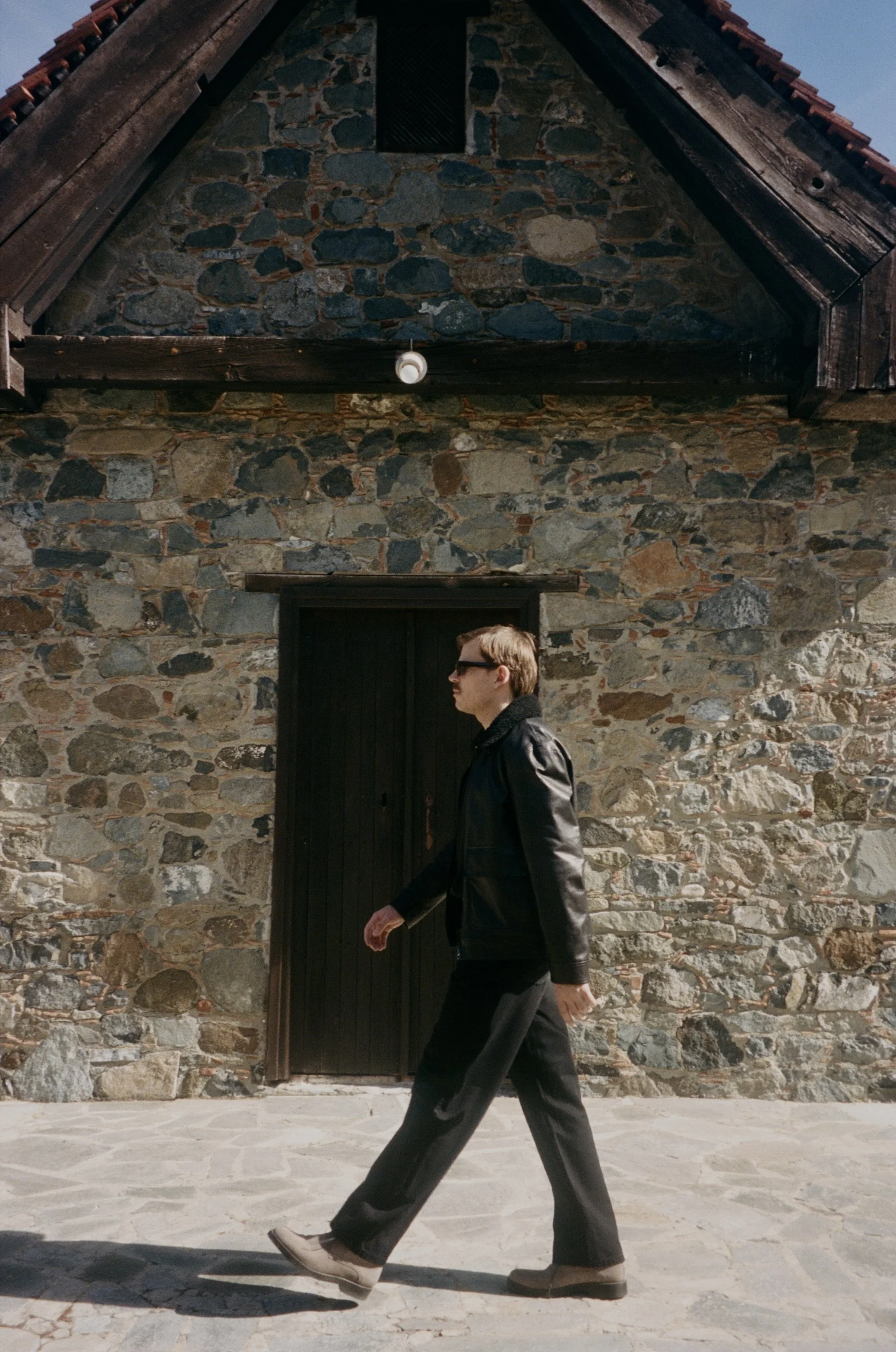 A man in dark clothing and sunglasses walks past a stone building with a black door and a sloped roof with exposed wooden beams.