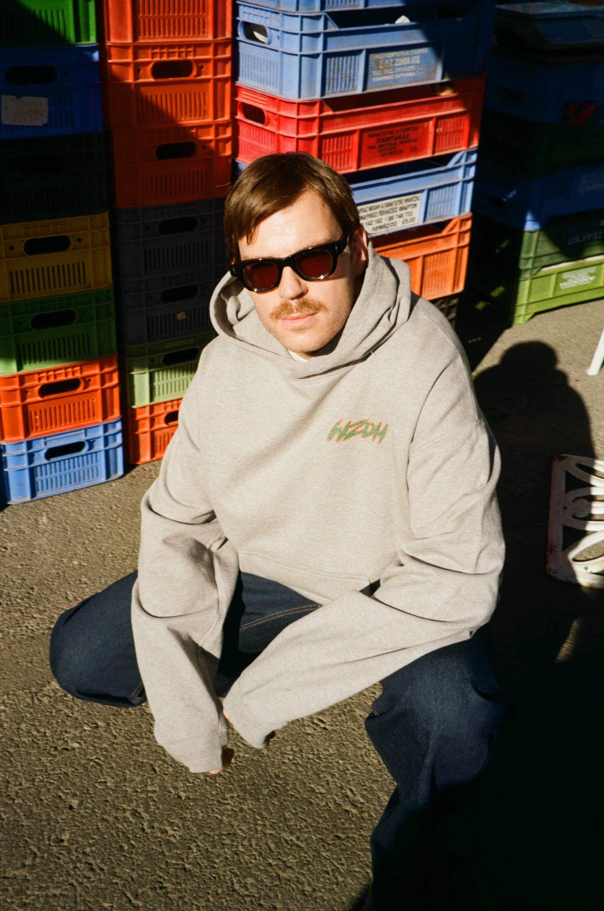 A man with brown hair, wearing dark sunglasses, a gray hoodie with a small logo, and dark jeans, is squatting on a street with stacks of colorful plastic crates behind him.