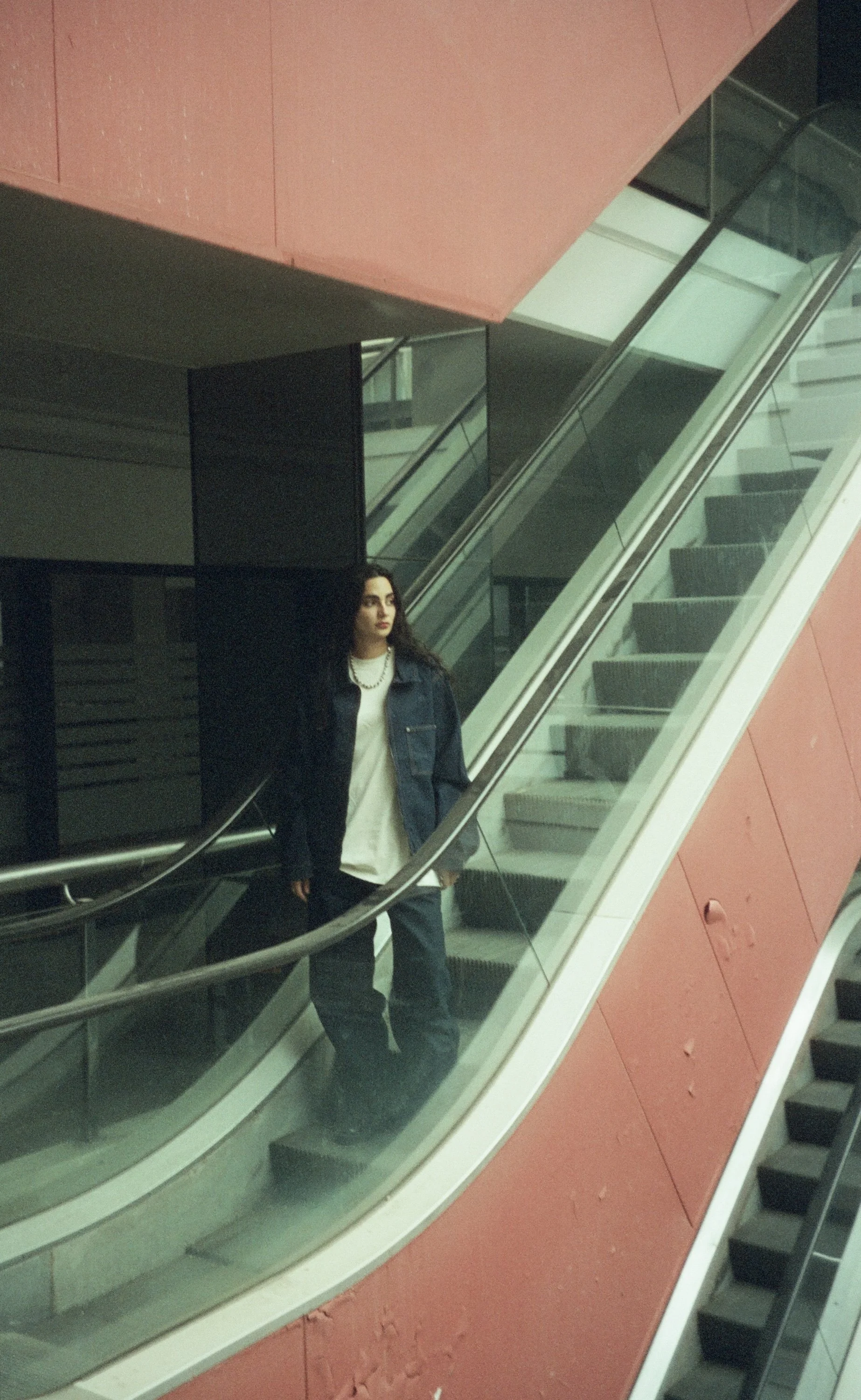 A young woman with long dark hair, wearing a denim jacket, white shirt, and black pants, stands on an escalator inside a modern building with pink walls, black panels, and glass railings.
