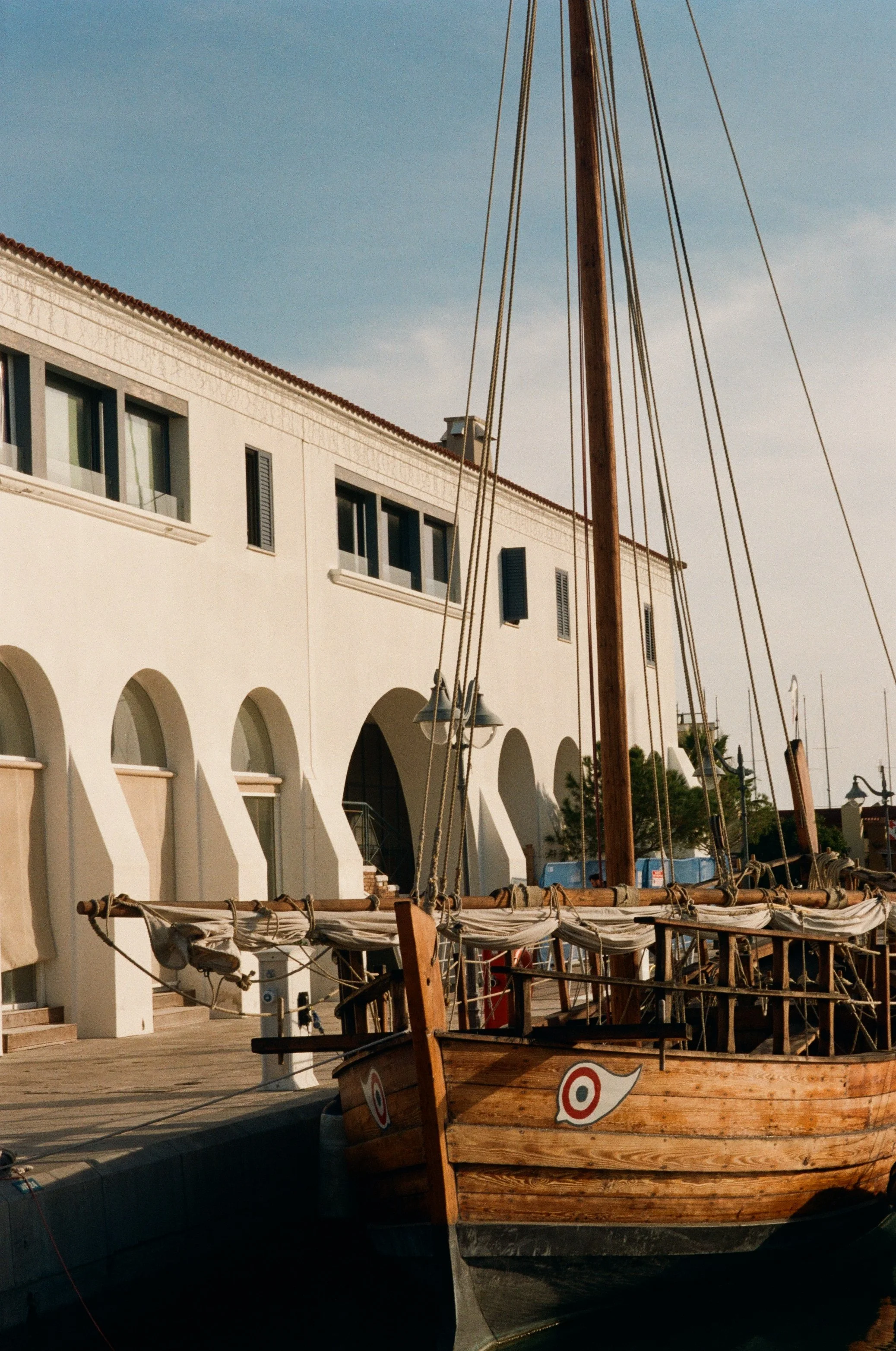 A wooden sailboat docked at a marina with a white building featuring arched windows in the background.