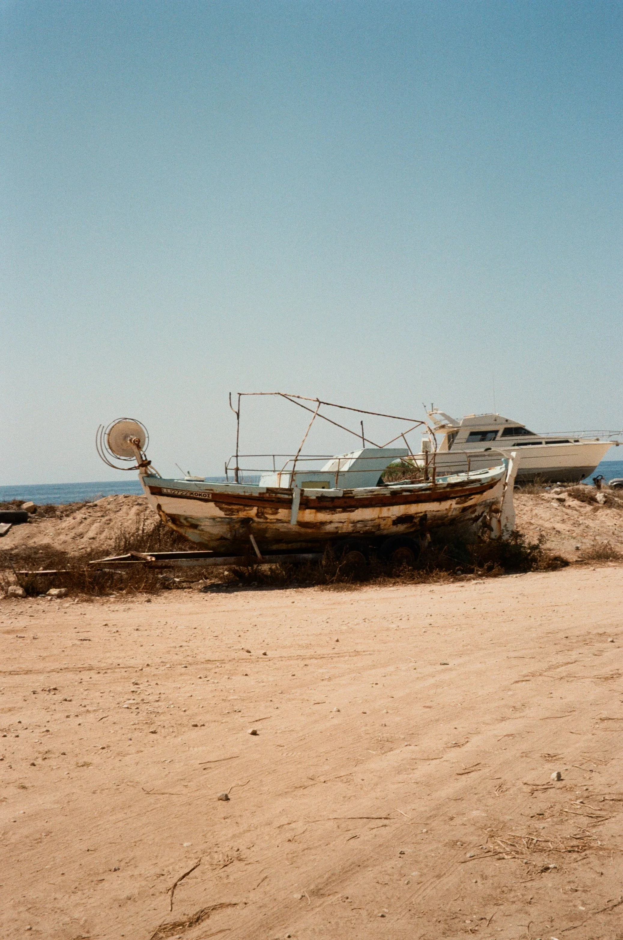 Two boats, one old and rusty on a trailer and one modern yacht, resting on rocky land near the beach under a clear blue sky.