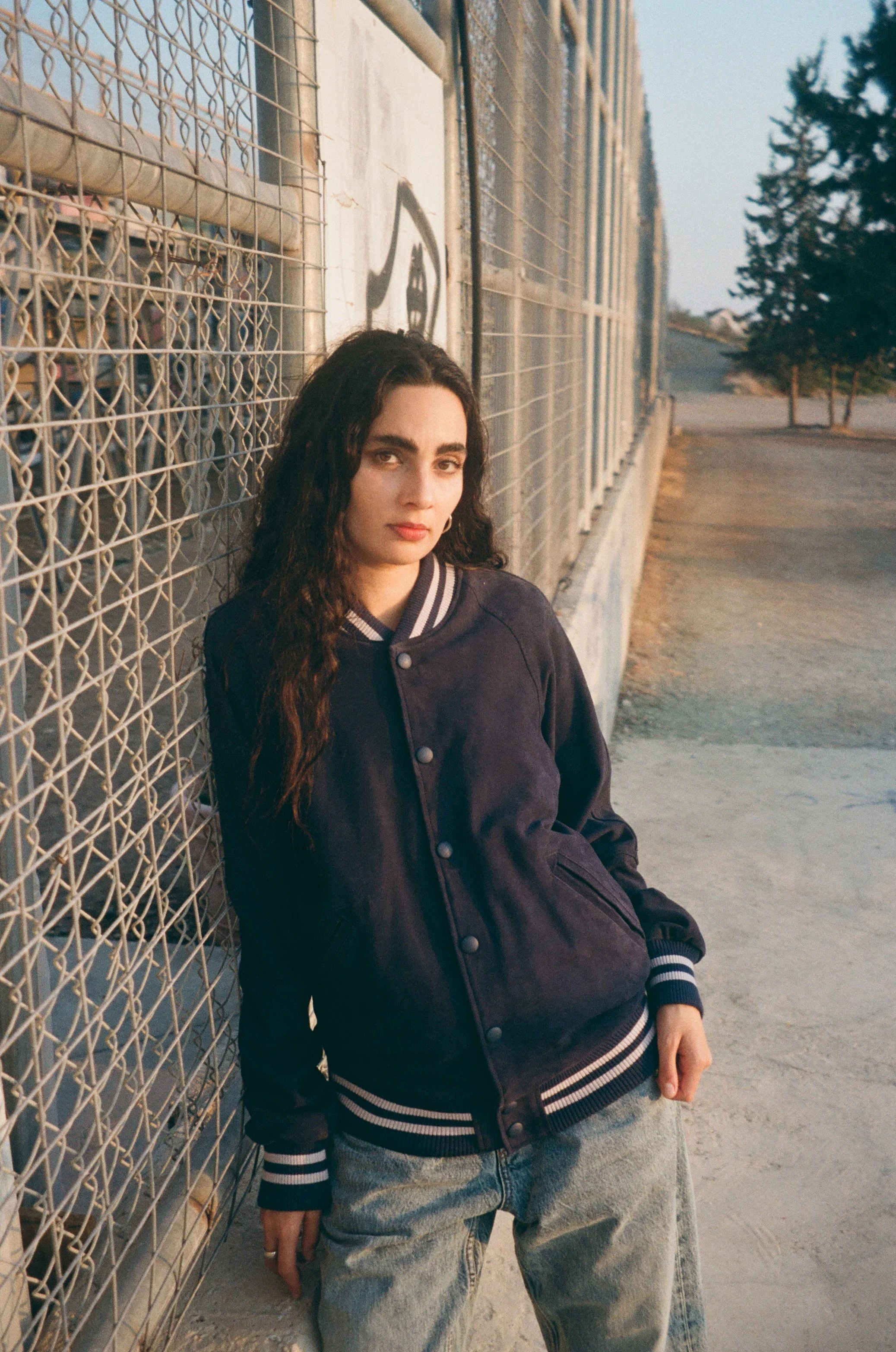 A young woman with long, dark wavy hair leaning against a chain-link fence, wearing a navy blue varsity jacket with white stripes and light blue jeans, in an outdoor setting during sunset.
