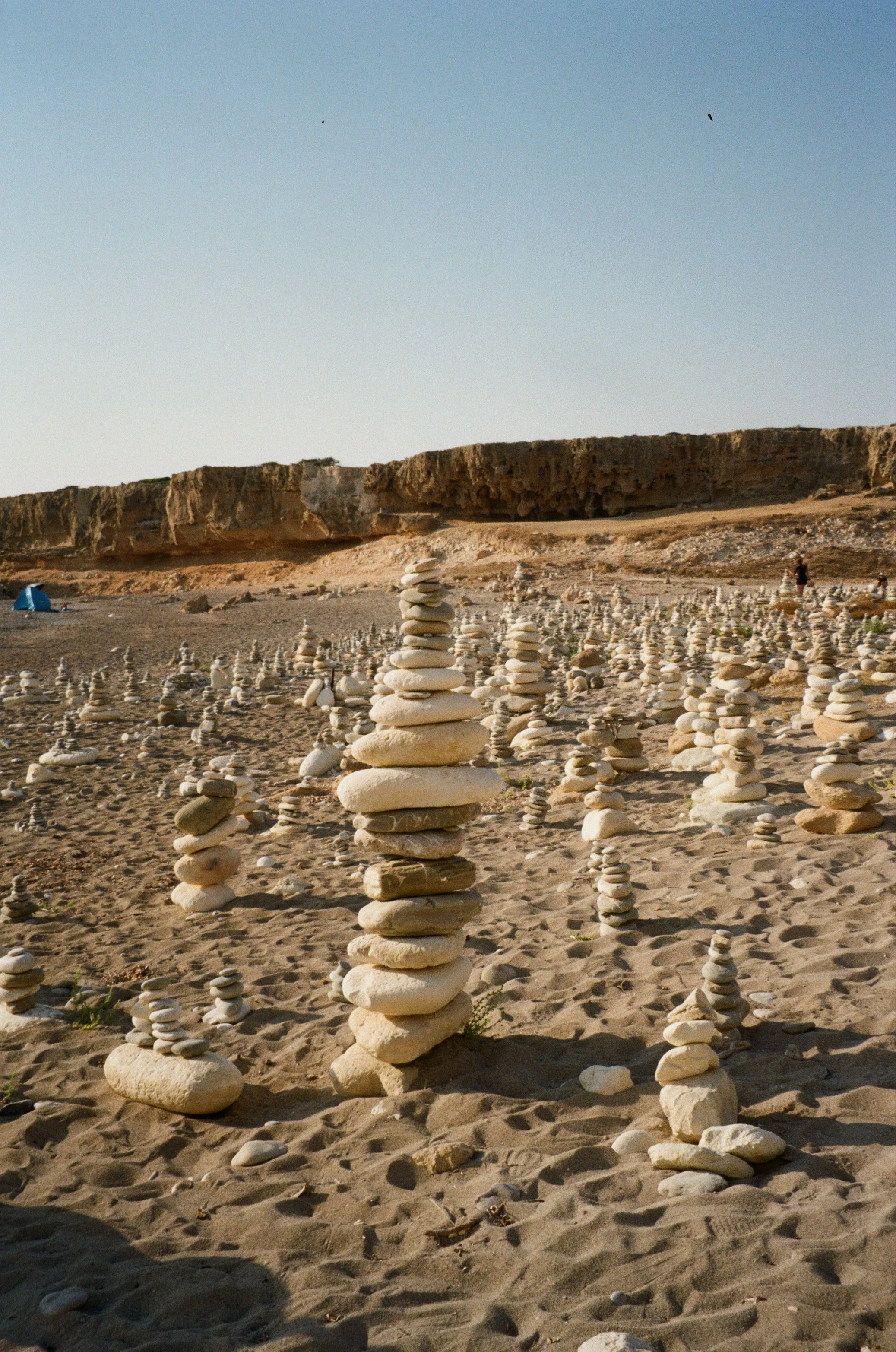 A desert beach with numerous stacks of smooth, flat stones arranged on the sand, with cliffs in the background and a clear sky above.