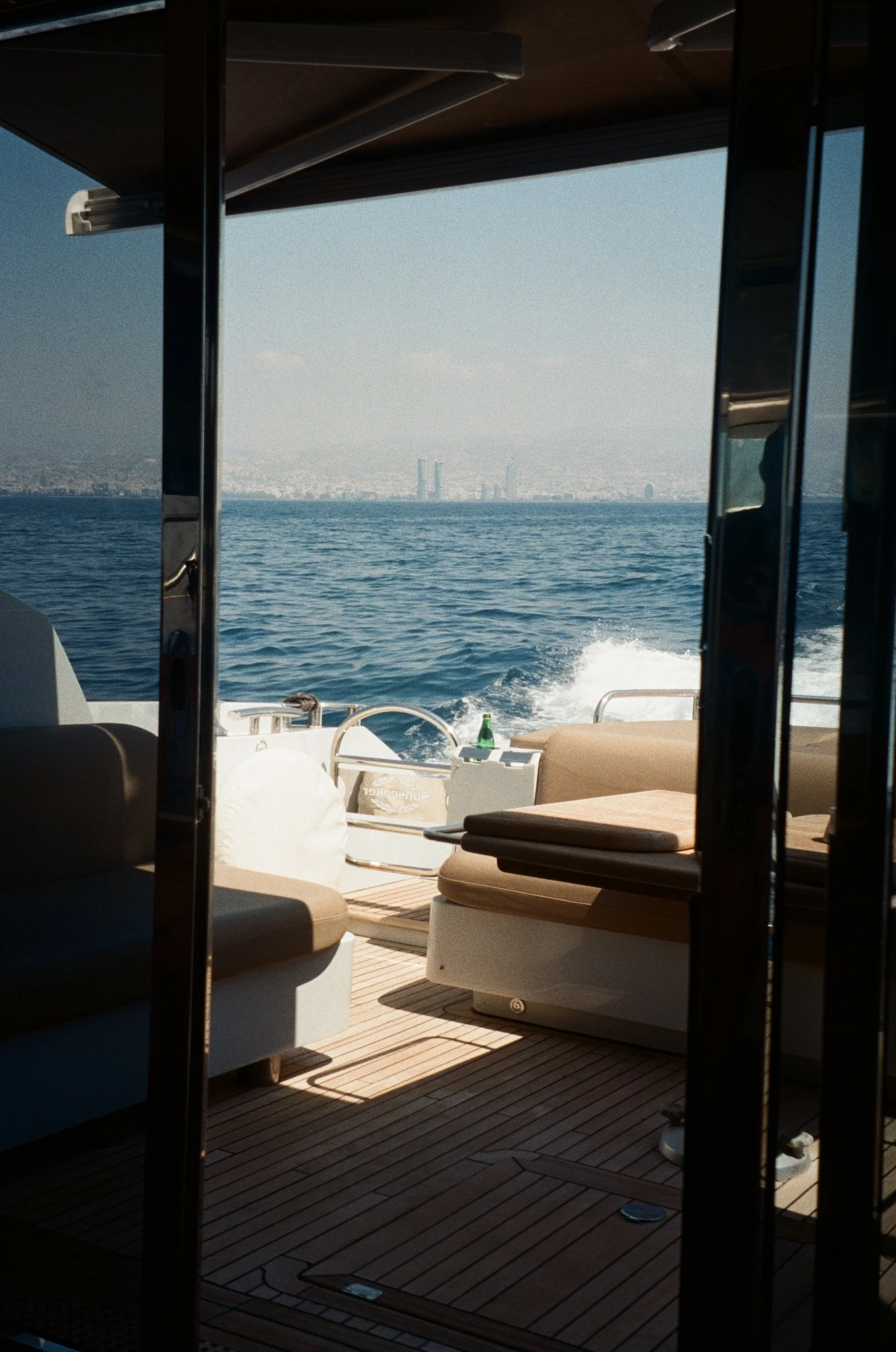 View of the ocean from inside a yacht, with outdoor furniture and a small table, and city skyline in the distance.