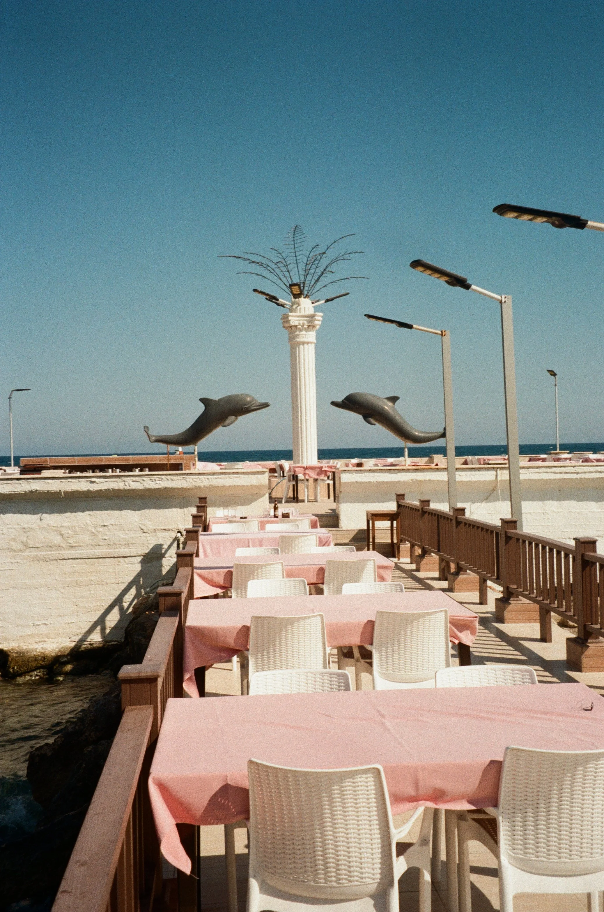 Outdoor seaside restaurant with pink tablecloth-covered tables, white chairs, dolphin sculptures, and a column topped with metal palm fronds, overlooking the ocean.