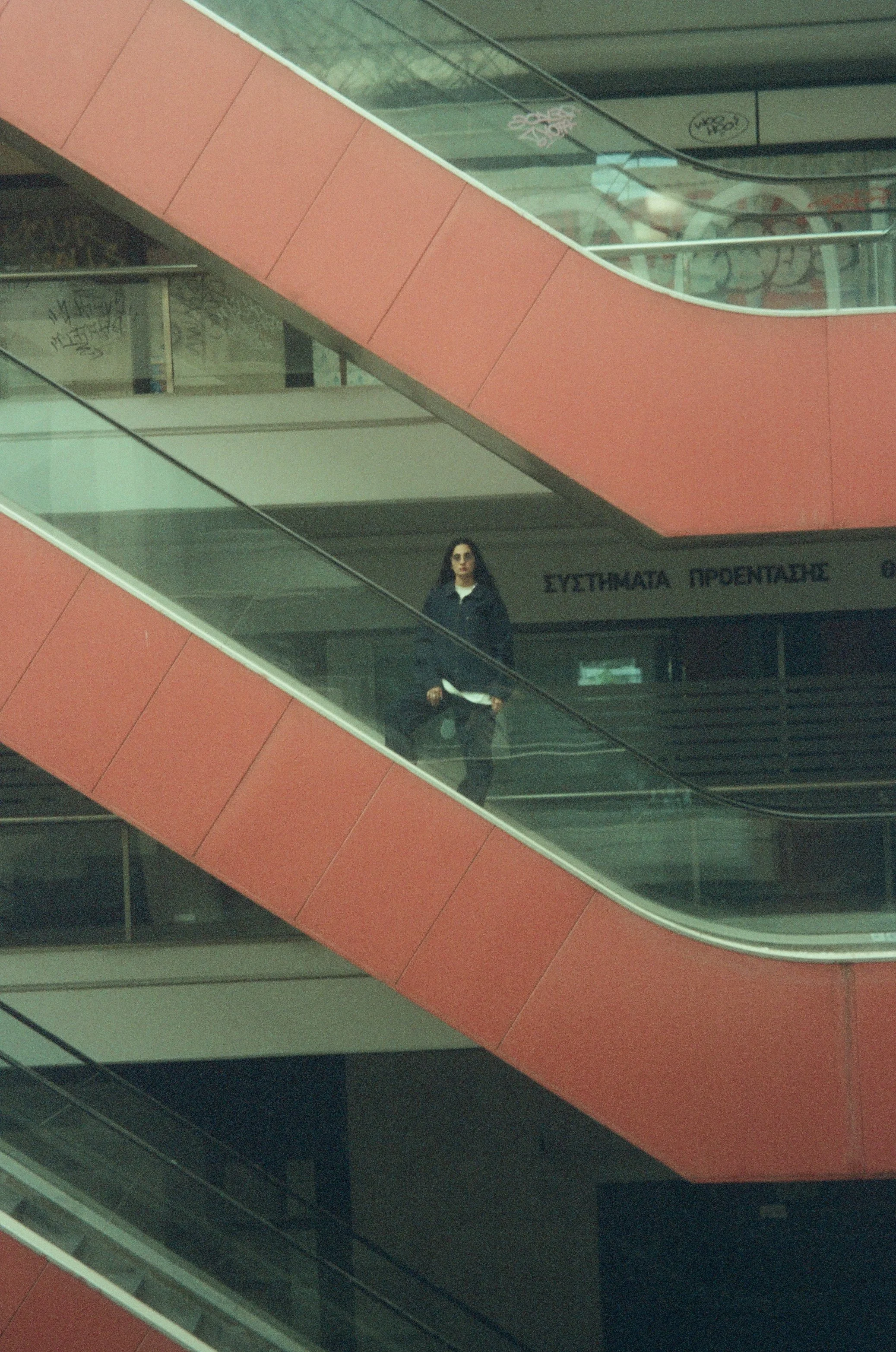 A woman standing on an escalator in a modern building, with red and glass railings and Greek lettering in the background.