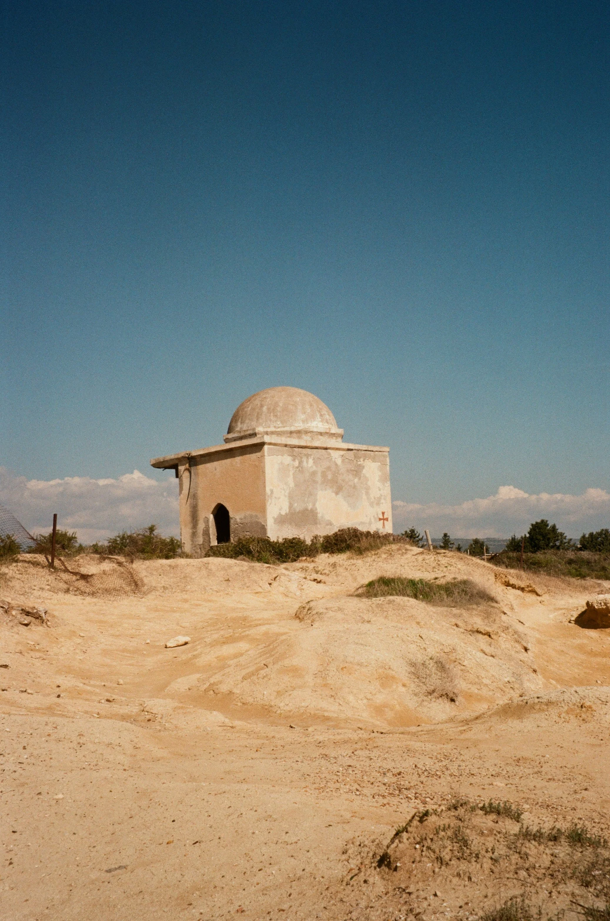 A small, weathered stone building with a rounded dome on top, situated in a dry, sandy landscape under a clear blue sky.