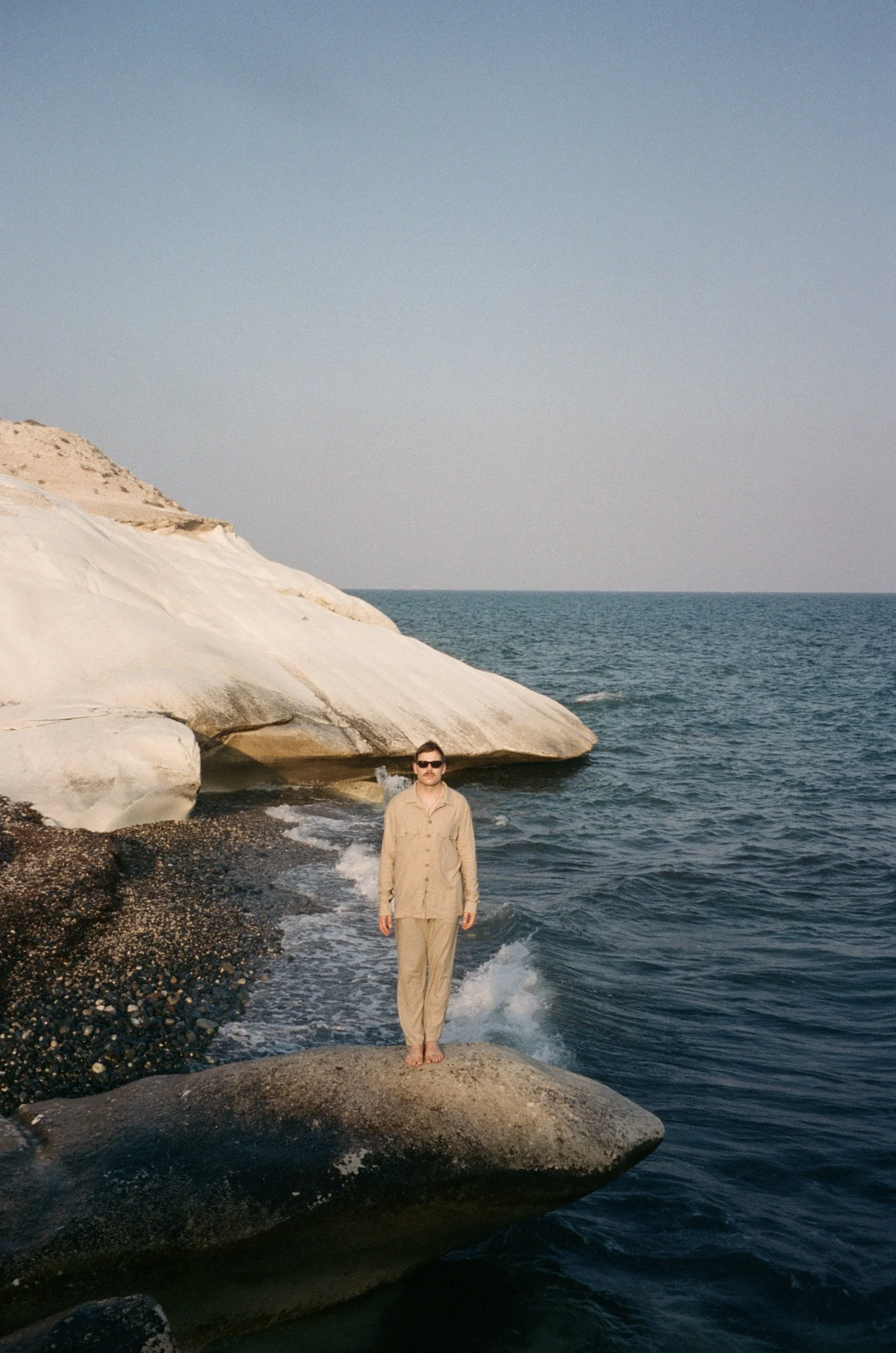 A person standing on a large rock at the edge of the ocean, with white cliffs in the background and a clear sky.