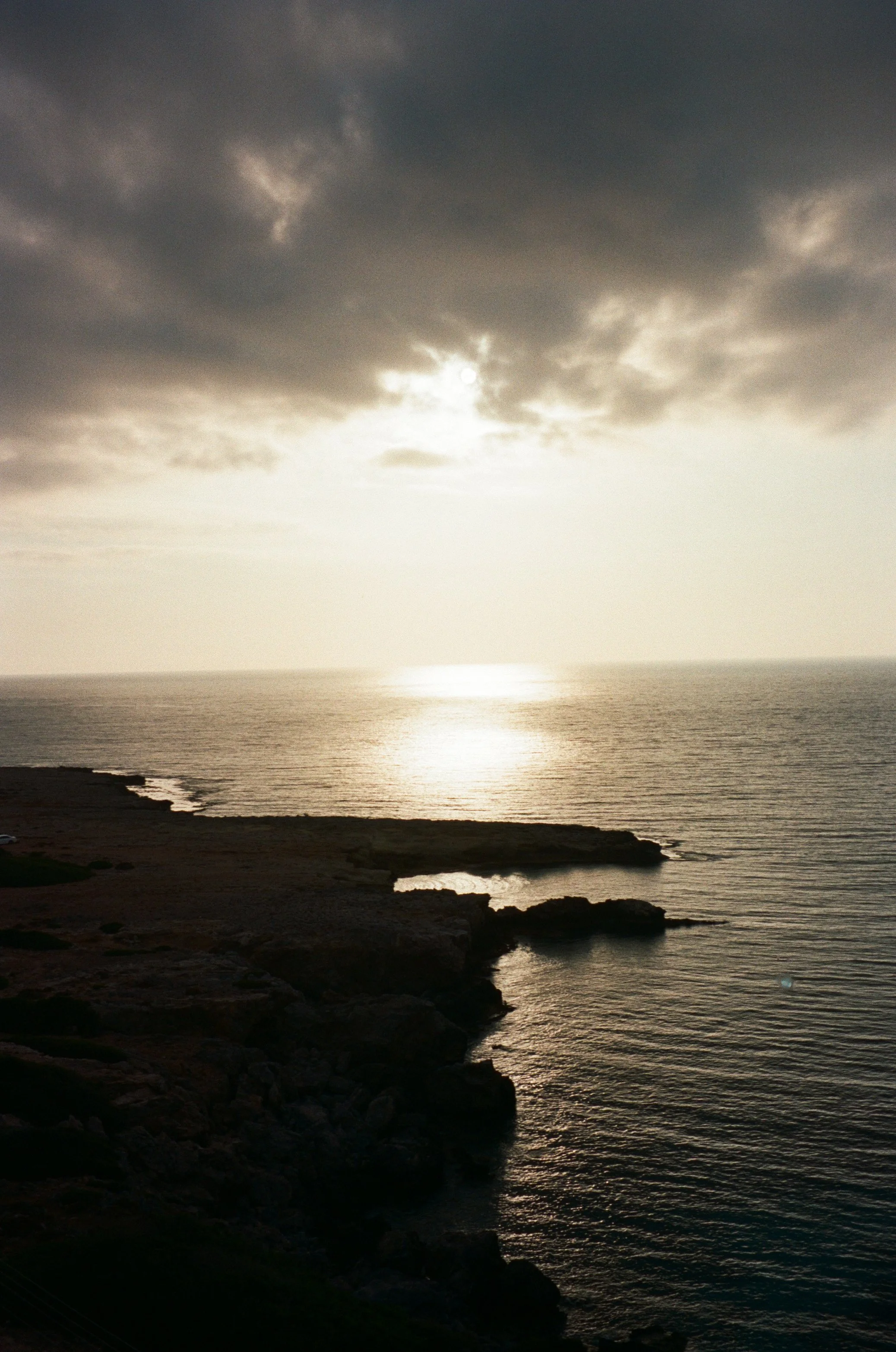 A photo of a coastal scene during sunset with dark clouds overhead, reflecting light on the water, and a rugged shoreline in the foreground.