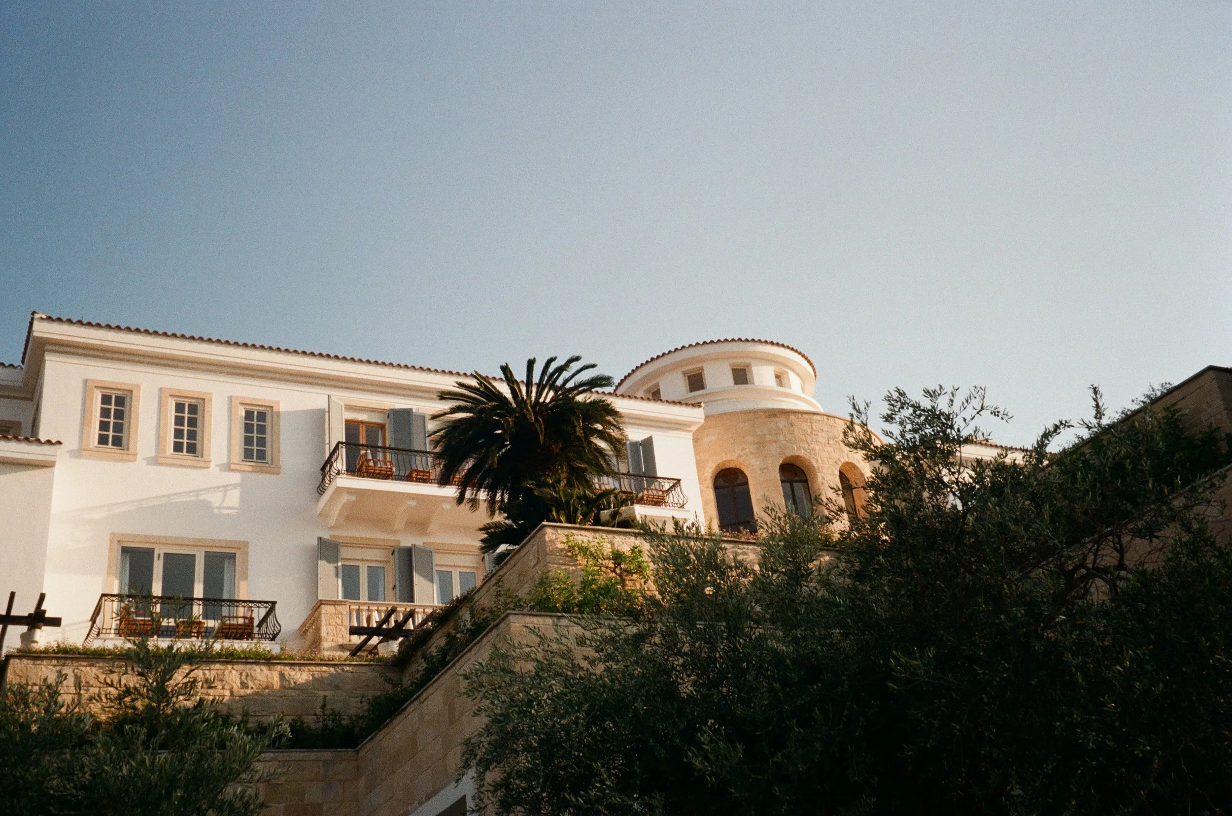 A large Mediterranean style house with white walls and red tile roof, surrounded by trees including a palm tree, with a clear sky overhead during sunset.