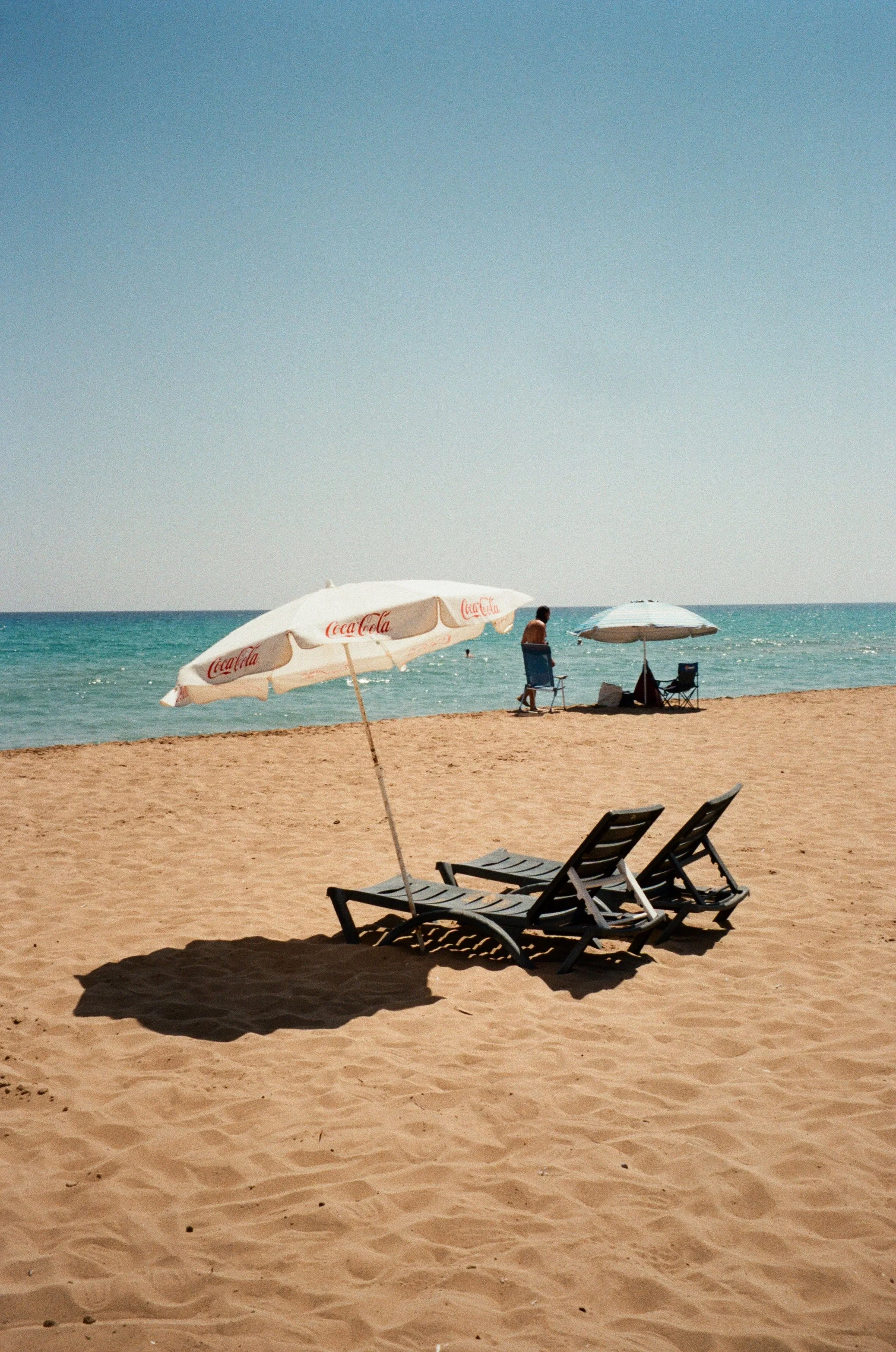 Empty beach with two lounge chairs and umbrellas, one branded with Coca-Cola, near the shoreline with a person standing in the background.