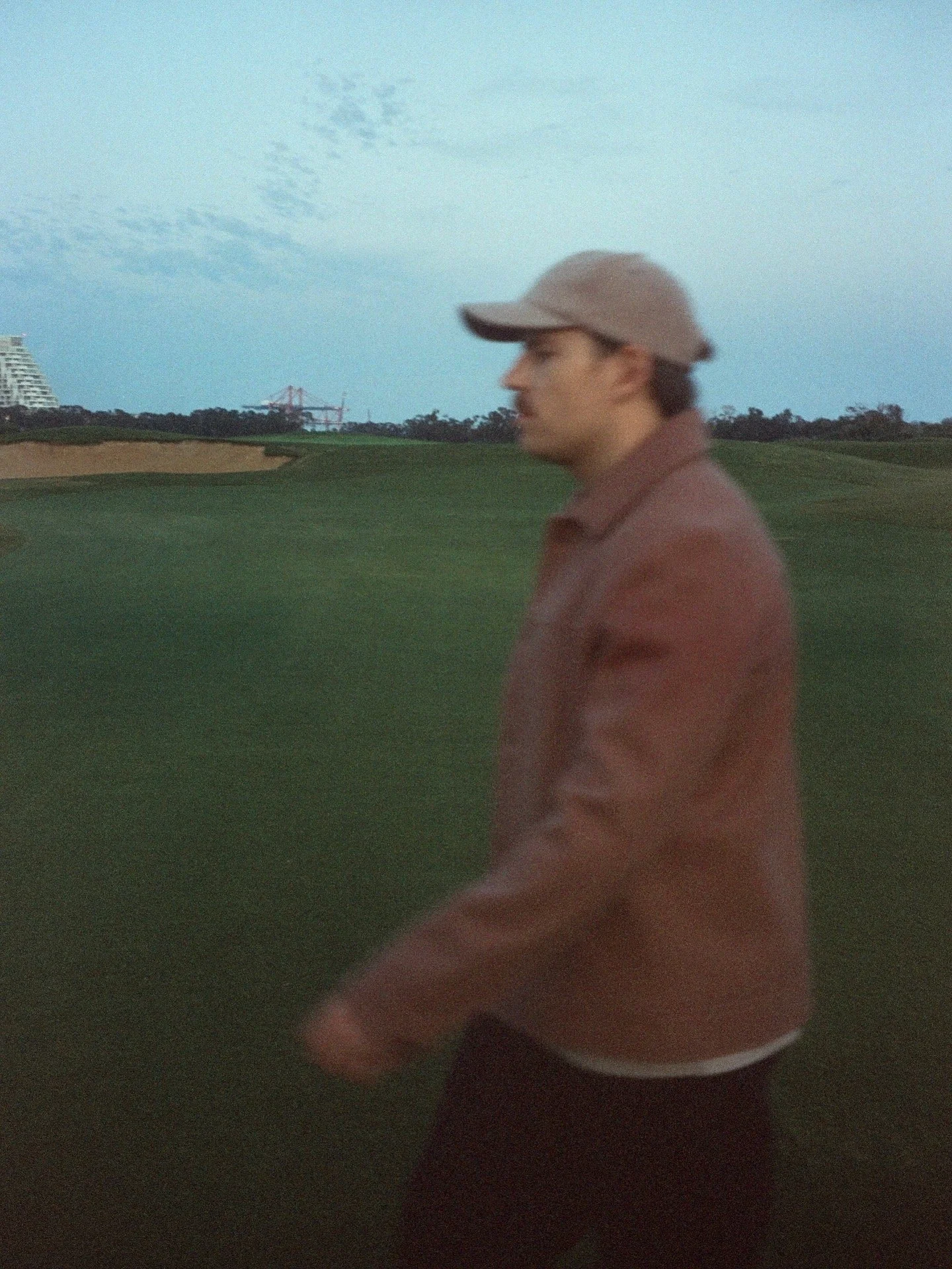 A man walking on a golf course with green grass, distant trees, and a blue sky with scattered clouds in the background, wearing a beige cap and brown jacket.