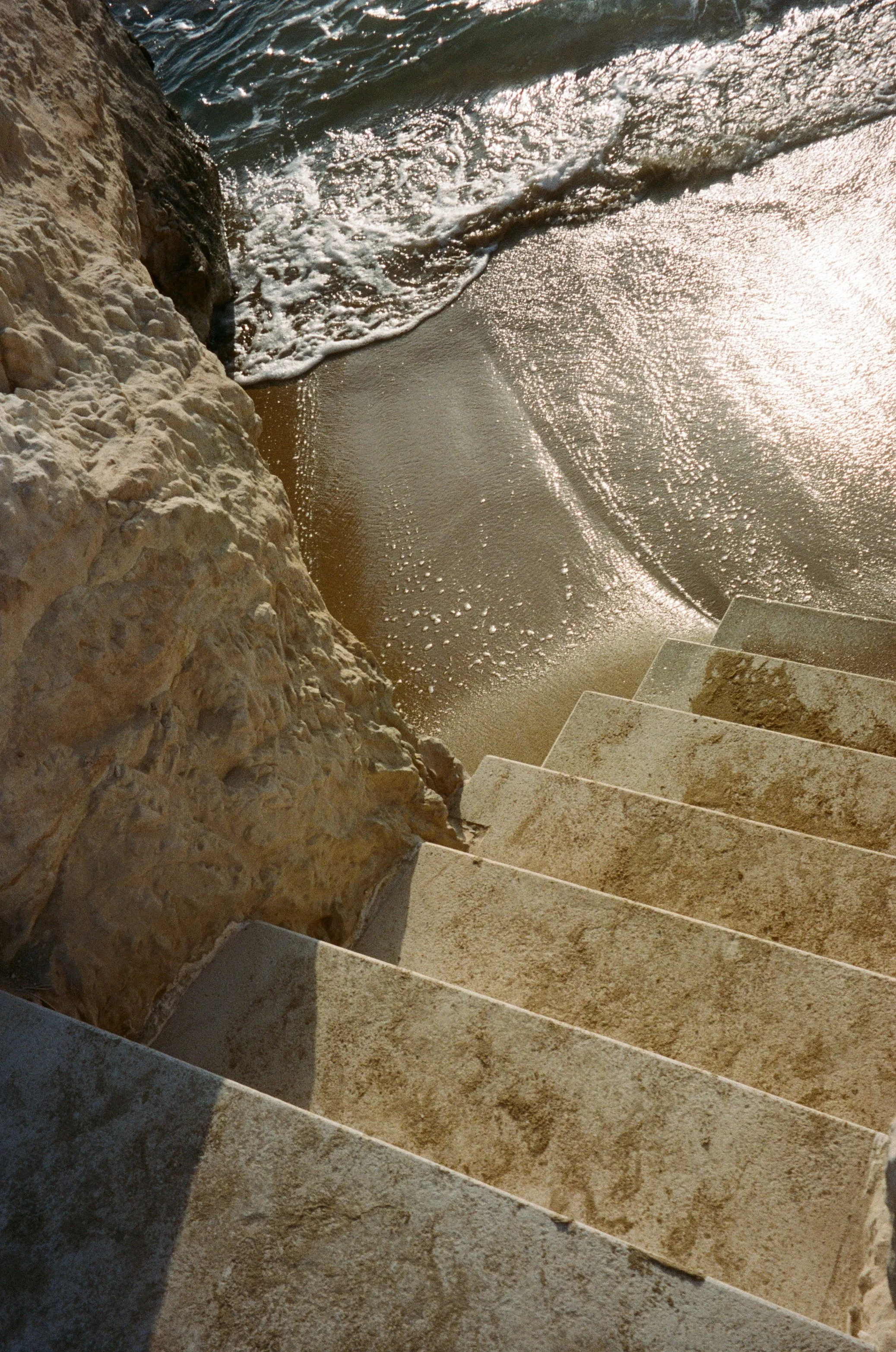 Concrete stairs next to a rocky wall leading down to the sandy shore, with waves gently lapping at the beach in the sunlight.