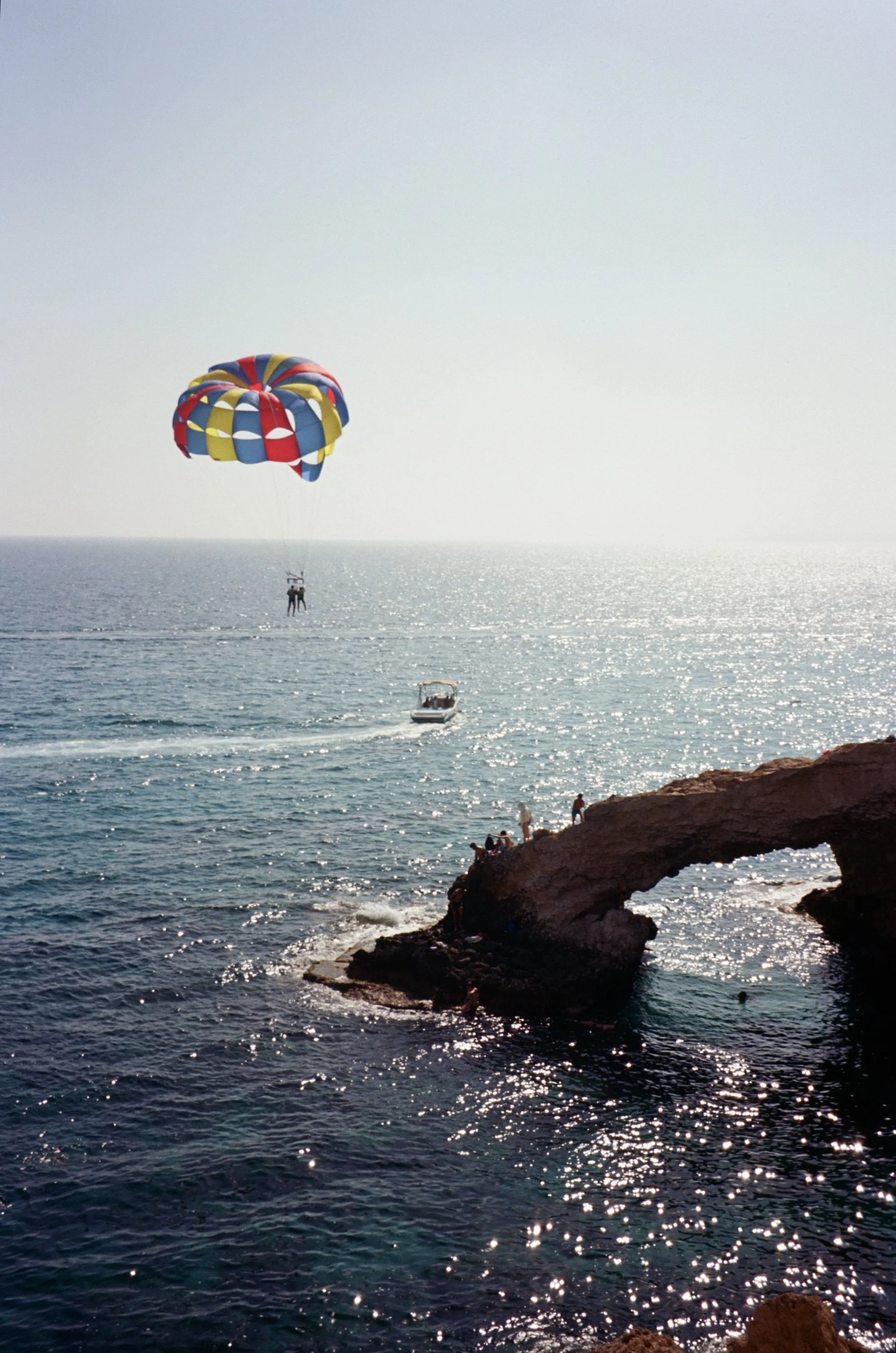 People parasailing near a rocky shoreline with a boat in the water and a natural stone arch formation